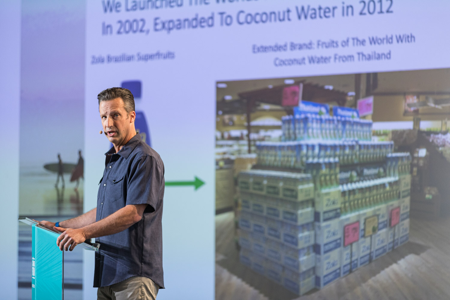 A man in a gray shirt stands at a podium beside a projected slide reading We Launched The World's First Packaged Acai Juice, In 2002, Expanded To Coconut Water in 2012 with a stacked retail display.