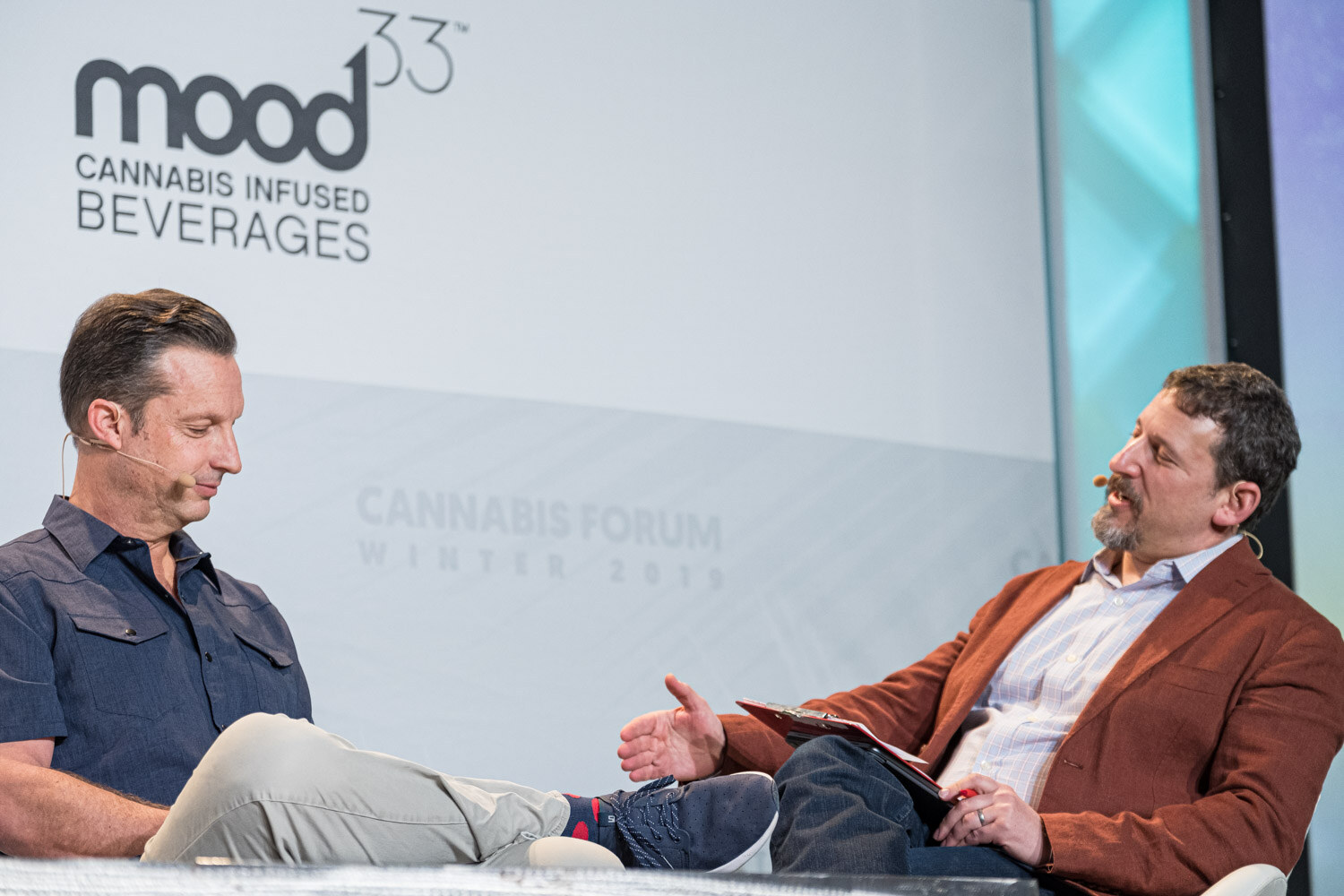 A man in a dark shirt looks down while a bearded man in a rust-colored jacket gestures toward him, with a Mood33 Cannabis Infused Beverages logo behind them.