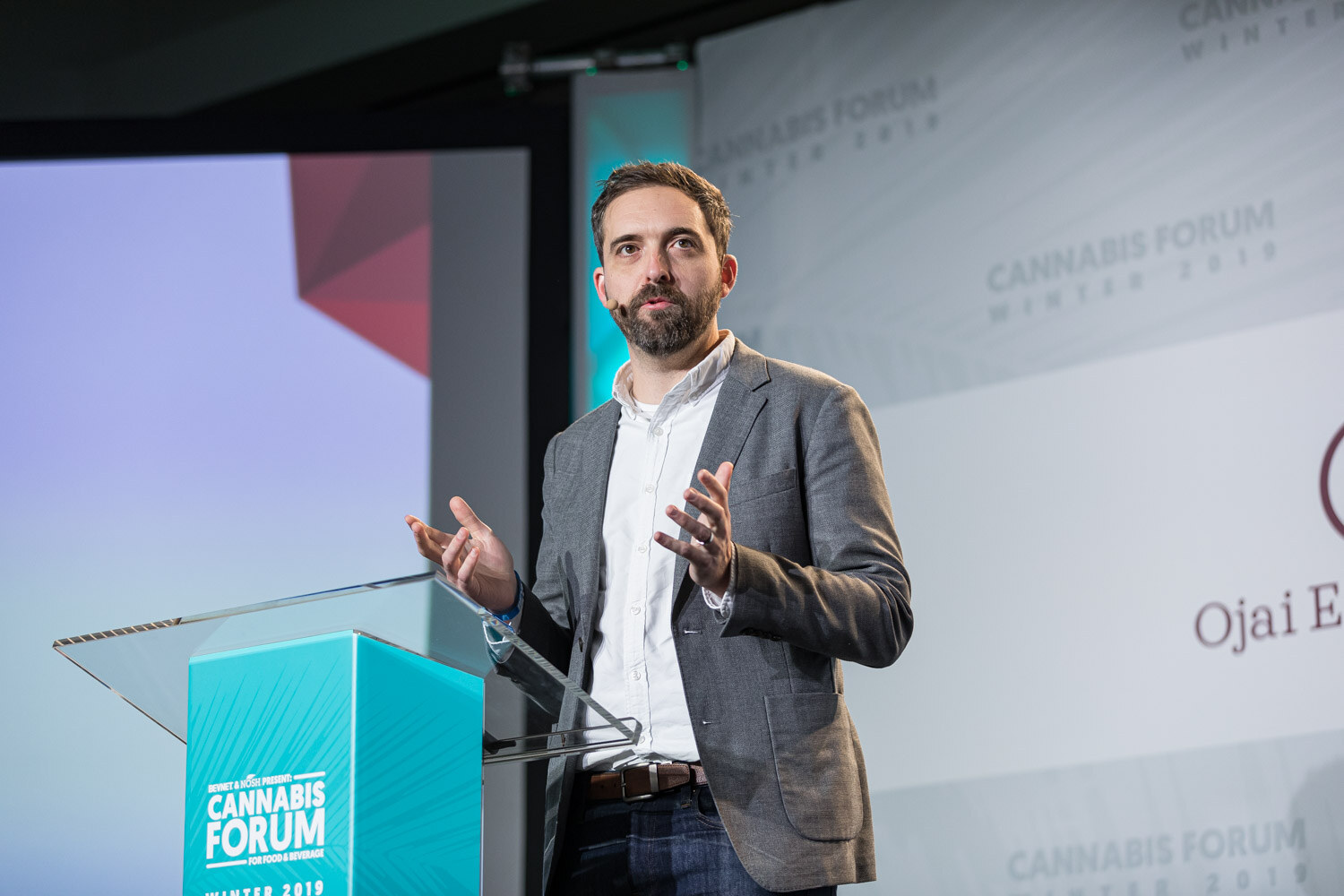 A bearded man in a gray blazer and white shirt holds both hands out at a teal podium, with a purple slide edge and a partial Ojai Energetics logo behind him.