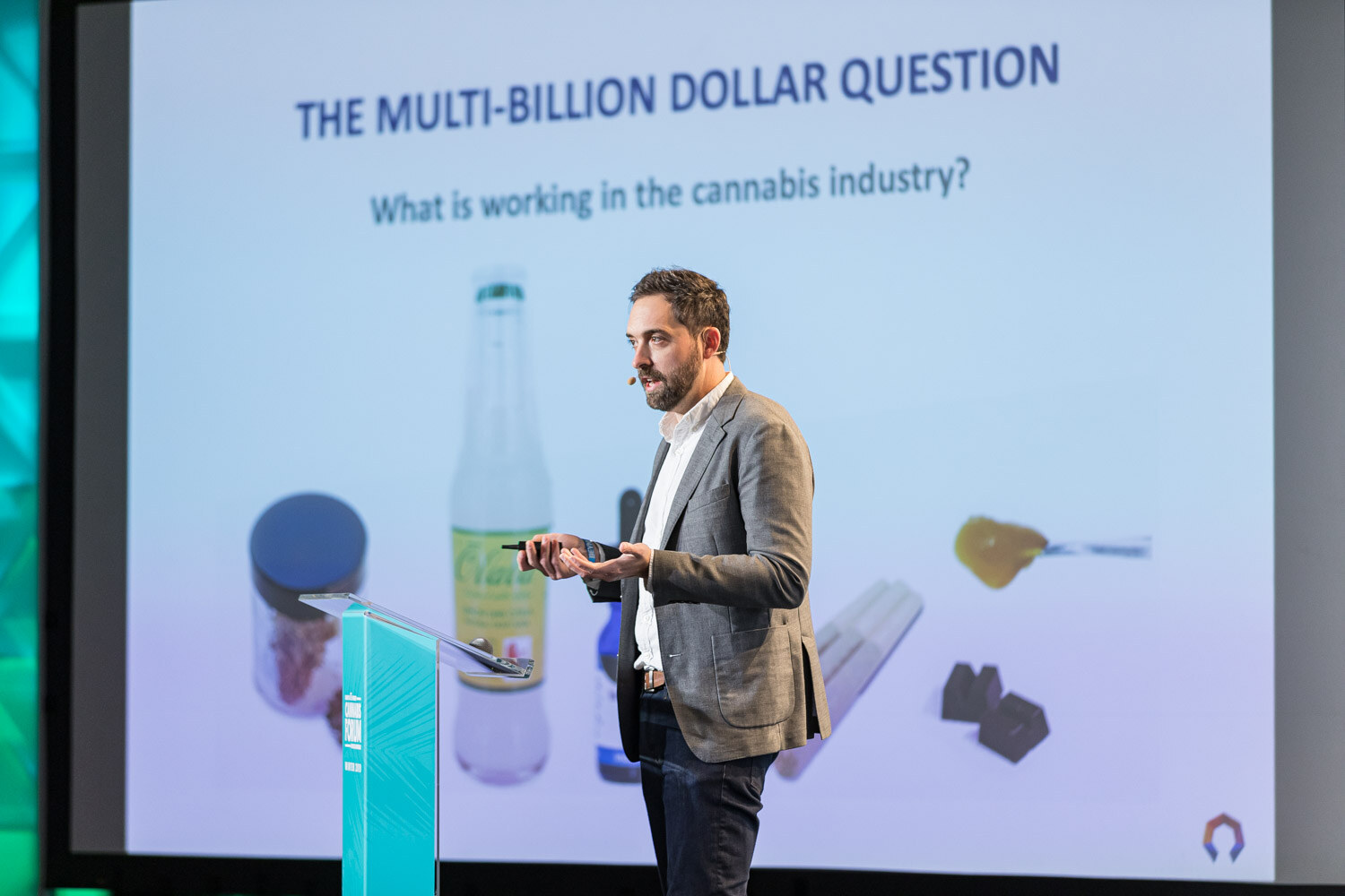 A man in a gray blazer stands at a podium in front of a slide reading The Multi-Billion Dollar Question with pictures of a glass drink bottle, a jar of flower, a dropper bottle, pre-rolls and chocolates.
