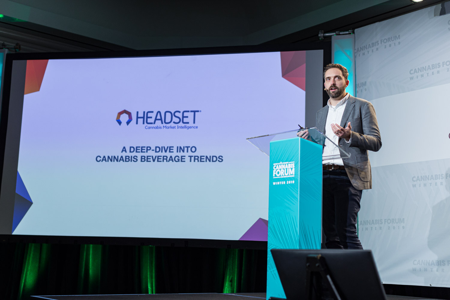 A man in a gray blazer stands at a podium beside a wide screen reading Headset Cannabis Market Intelligence, A Deep-Dive Into Cannabis Beverage Trends.