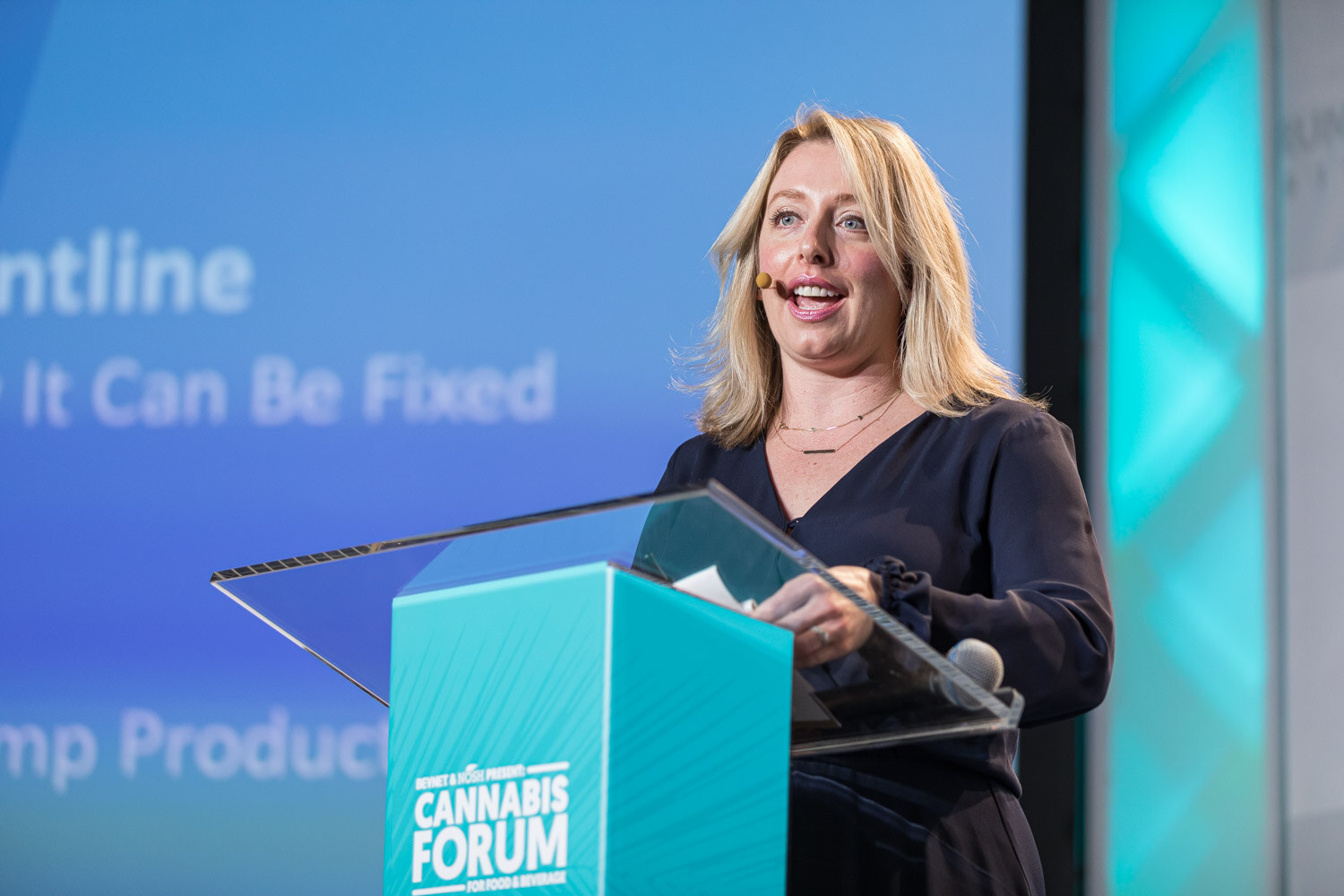 A blond woman in a navy top speaks smiling at a teal podium, with the slide text Frontline and It Can Be Fixed behind her.