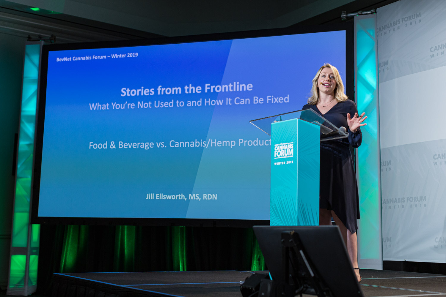 A woman in a navy dress stands at a teal podium at the right of a large blue slide titled Stories from the Frontline: What You're Not Used to and How It Can Be Fixed.