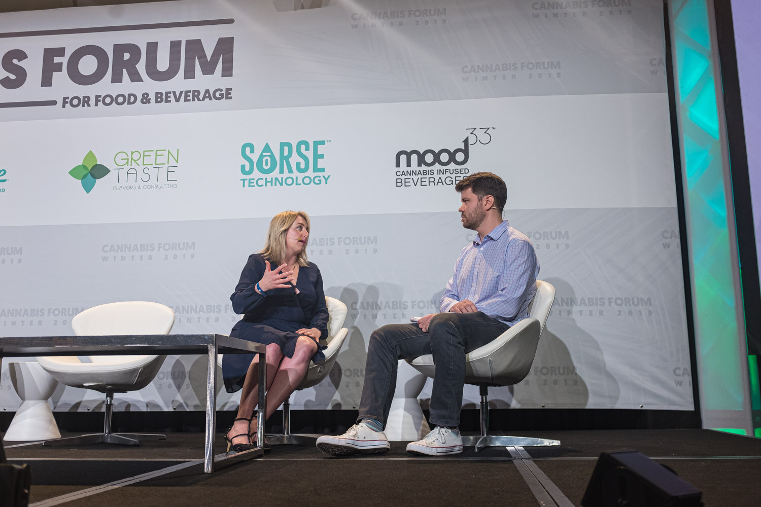 A woman in a navy dress and a man in a blue checked shirt and white sneakers sit in white lounge chairs on stage, in front of the Green Taste, Sorse and Mood33 logos.
