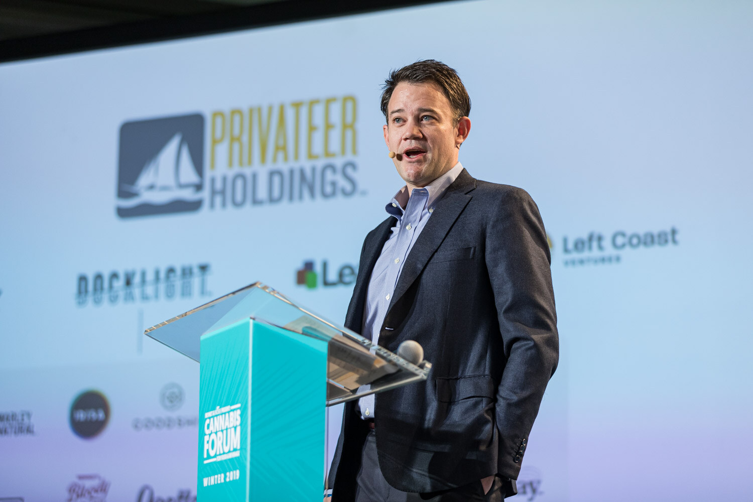 A man in a dark blazer stands at a podium with a hand in his pocket, in front of a slide with the Privateer Holdings logo and Docklight, Left Coast Ventures, Leafly and other logos.