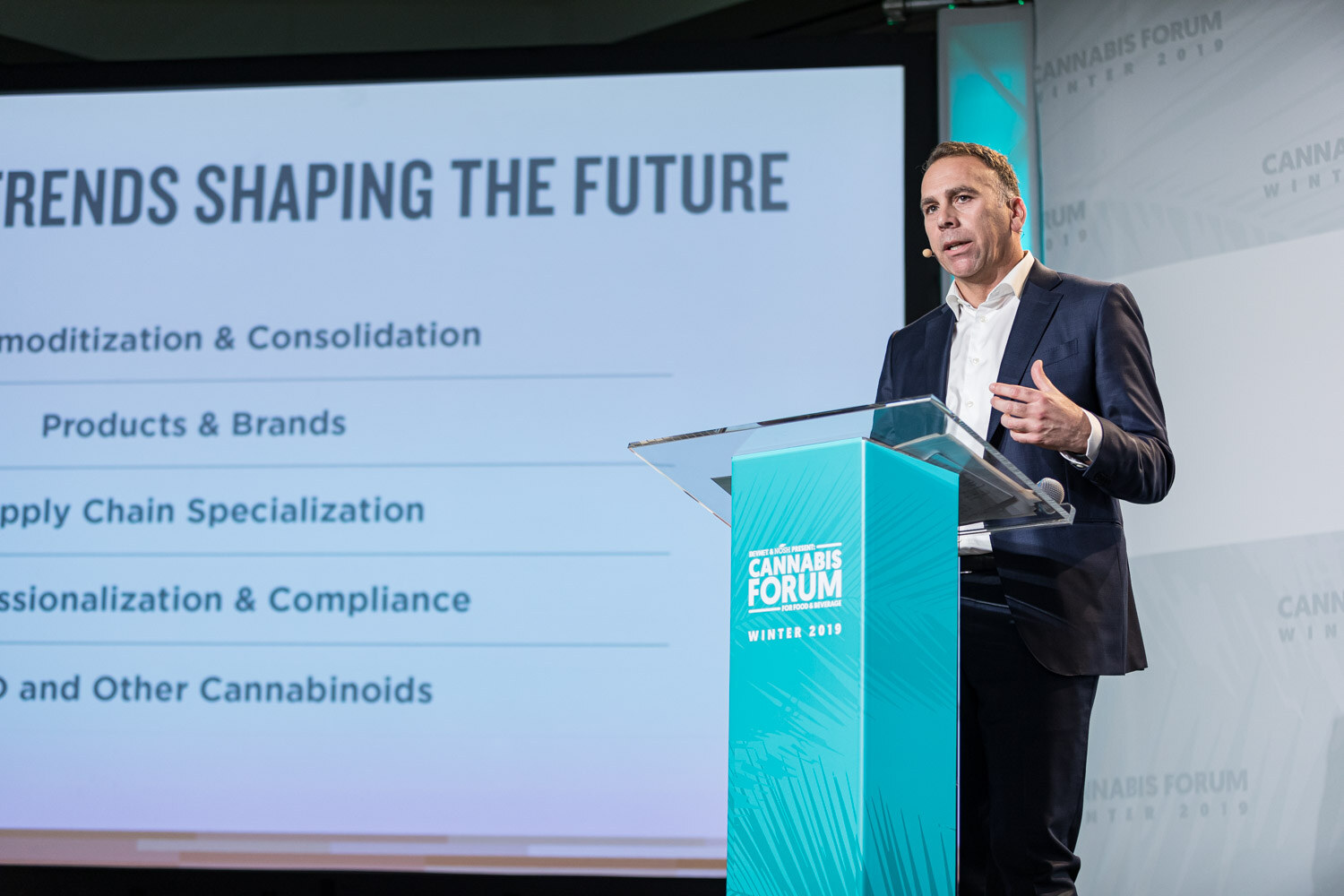 A man in a navy suit gestures at the podium beside a slide headed Trends Shaping The Future that lists commoditization and consolidation, products and brands, supply chain specialization, professionalization and compliance, and CBD and other cannabinoids.