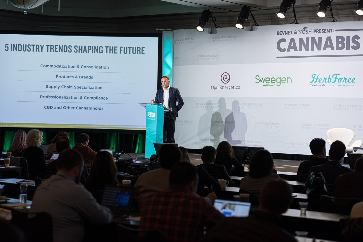 A wide view from the audience shows a speaker at the podium beside a large slide titled 5 Industry Trends Shaping The Future, with attendees and laptops in the foreground.