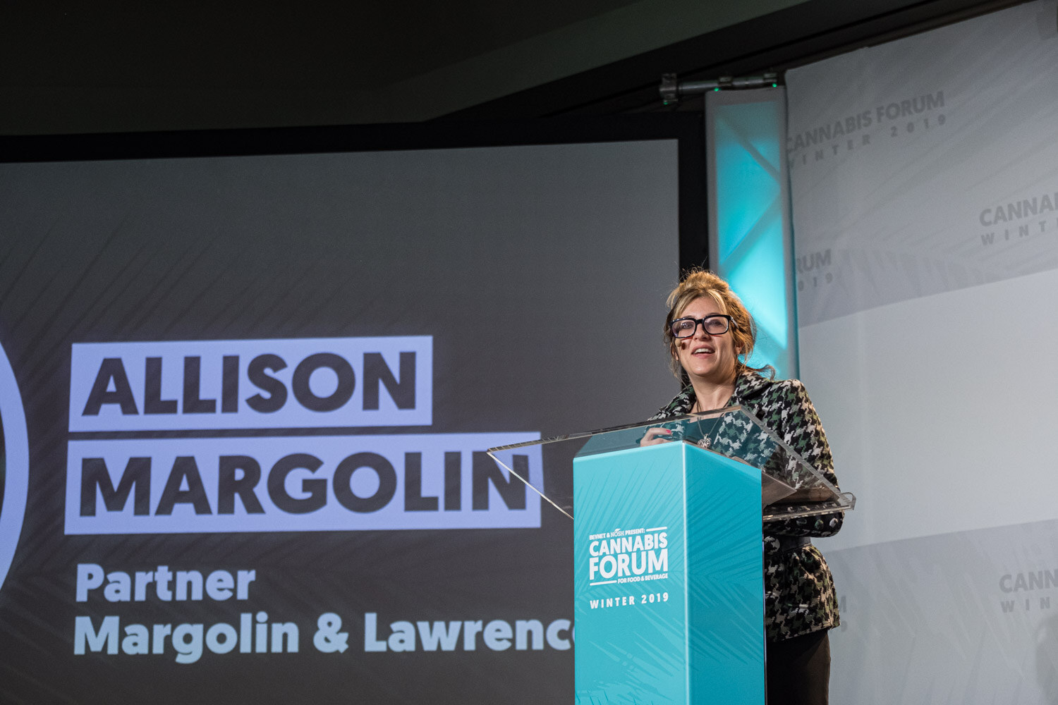 A woman in glasses and a houndstooth blazer stands at the podium beside a dark screen reading Allison Margolin, Partner, Margolin & Lawrence.