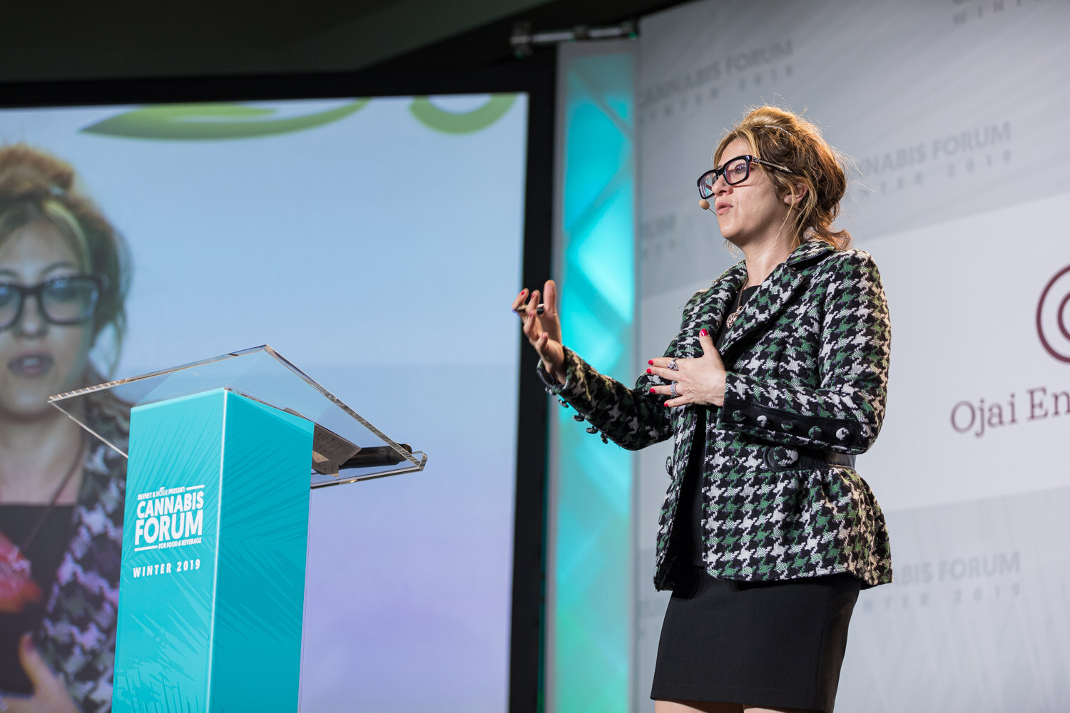 A woman in a houndstooth blazer and glasses gestures on stage, with her image enlarged on the screen behind her and an Ojai Energetics banner at right.