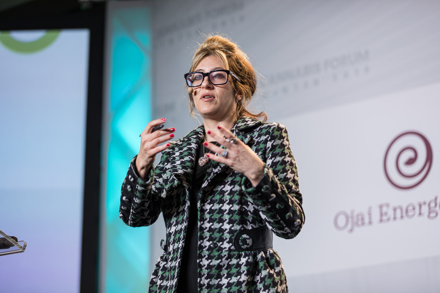 A close view of a woman in glasses and a houndstooth blazer speaking with both hands raised, with a headset microphone and an Ojai Energetics logo behind her.