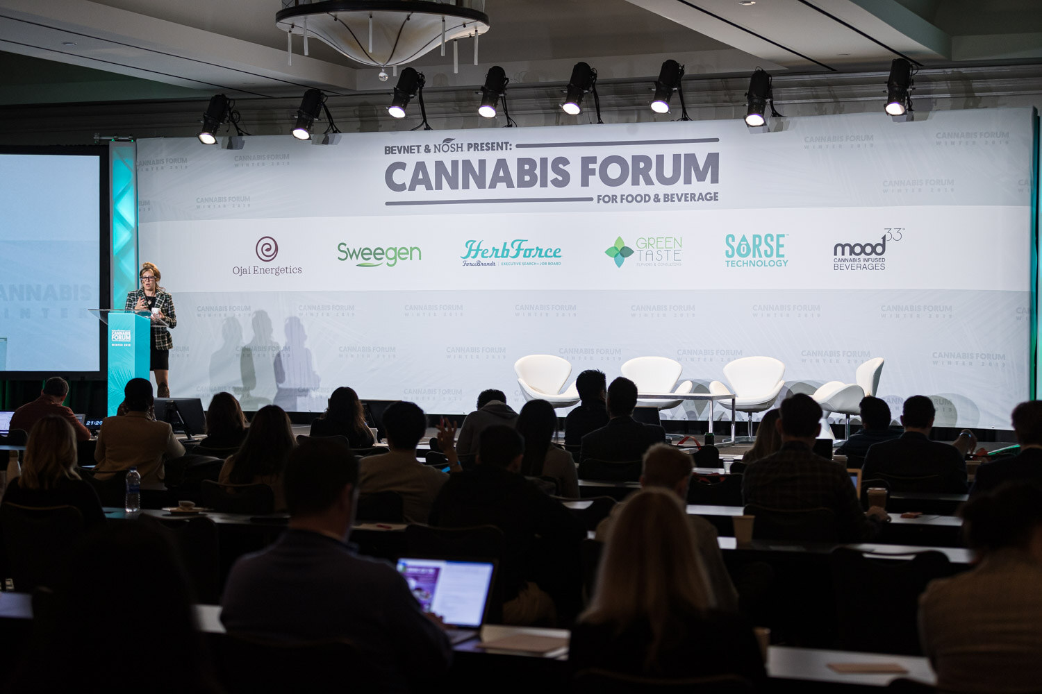 A wide ballroom view shows seated attendees in the foreground and a speaker at the podium in front of the Cannabis Forum backdrop with sponsor logos.