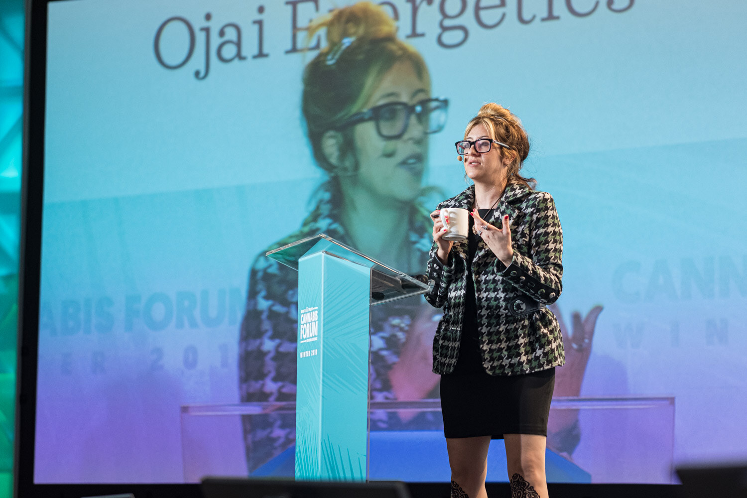 A woman in a houndstooth blazer, glasses and tall boots stands beside a teal podium holding a white mug, with a large Ojai Energetics screen and her enlarged image behind her.