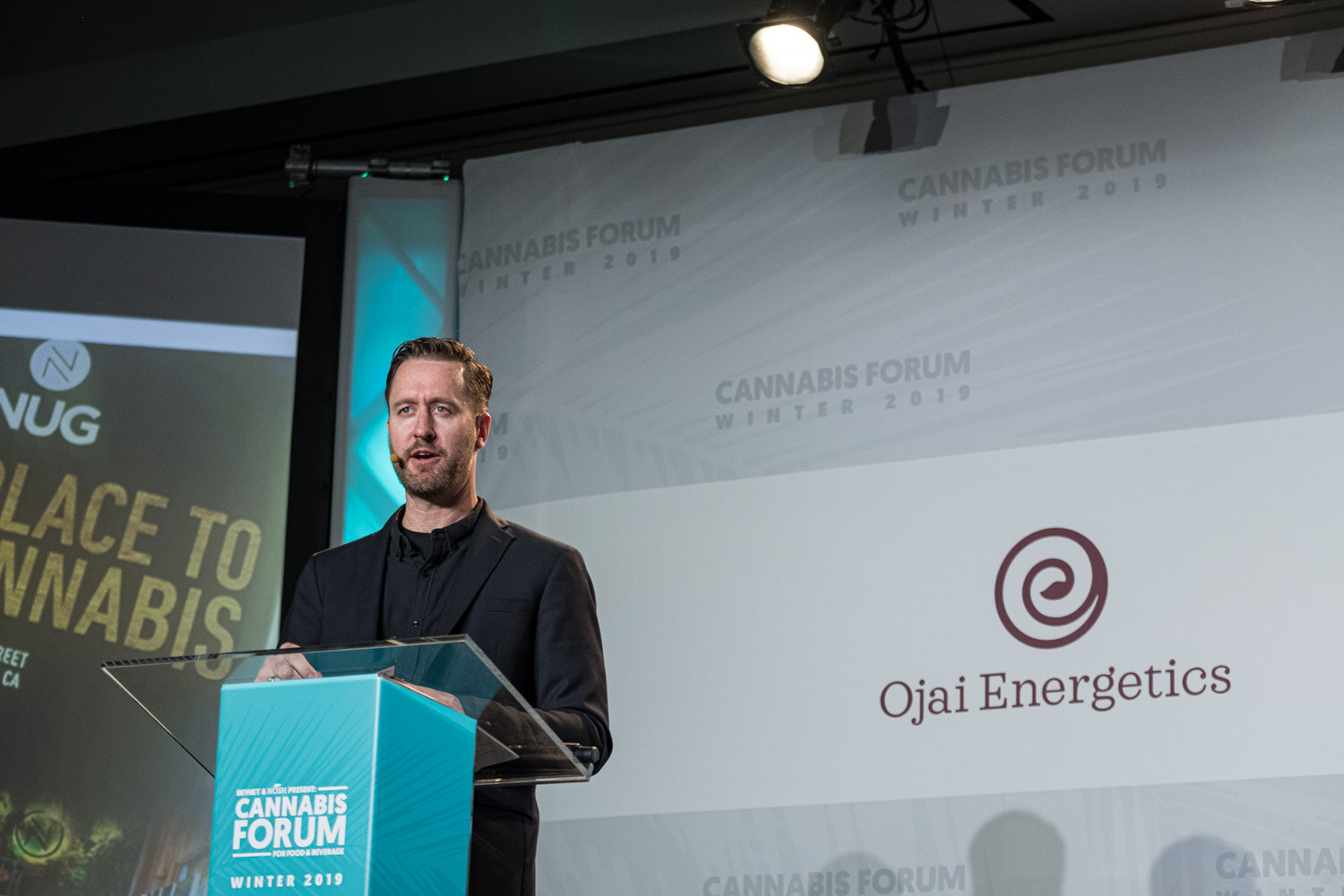 A man in a dark jacket speaks at a glass-topped podium with a NUG slide partly visible to the left and an Ojai Energetics logo at right.