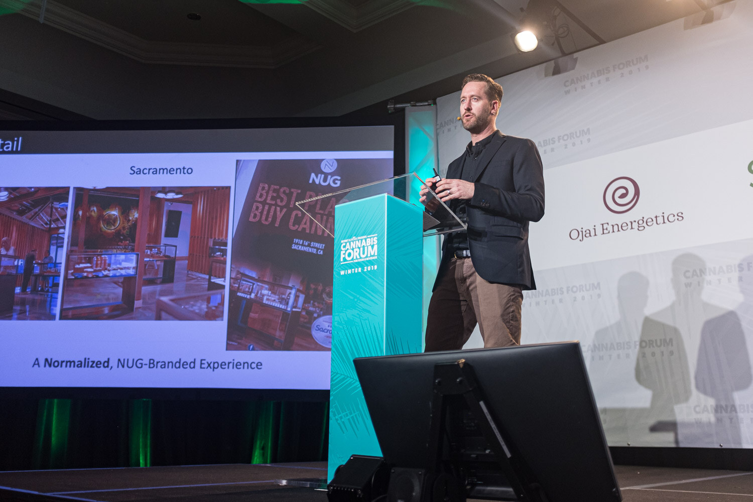 A man in a dark jacket stands at a teal podium beside a slide of a Sacramento store and NUG Best Place to Buy Cannabis cover, with an Ojai Energetics logo at right.