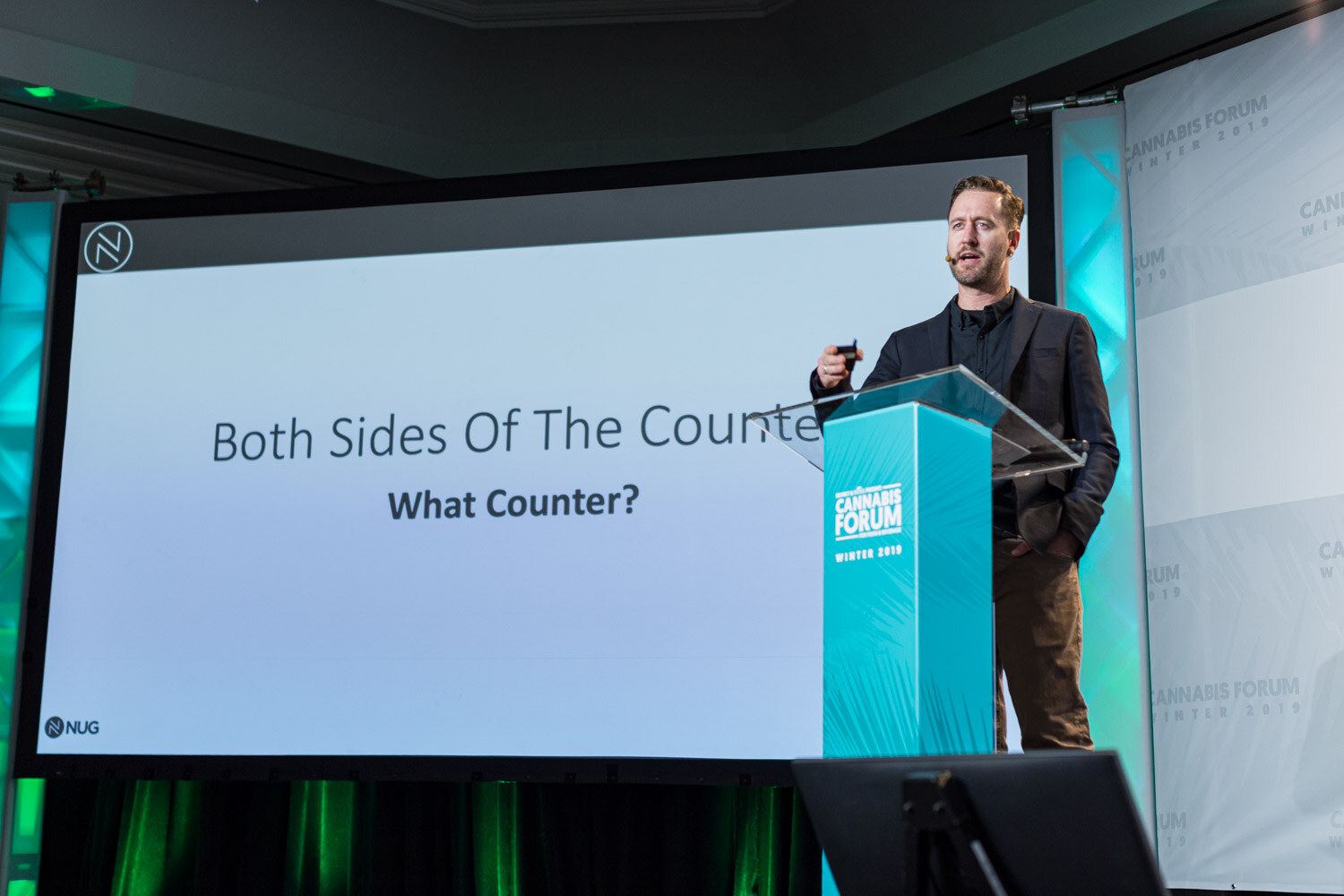 A man in a dark blazer stands beside a teal podium next to a projected slide reading Both Sides Of The Counter, What Counter?