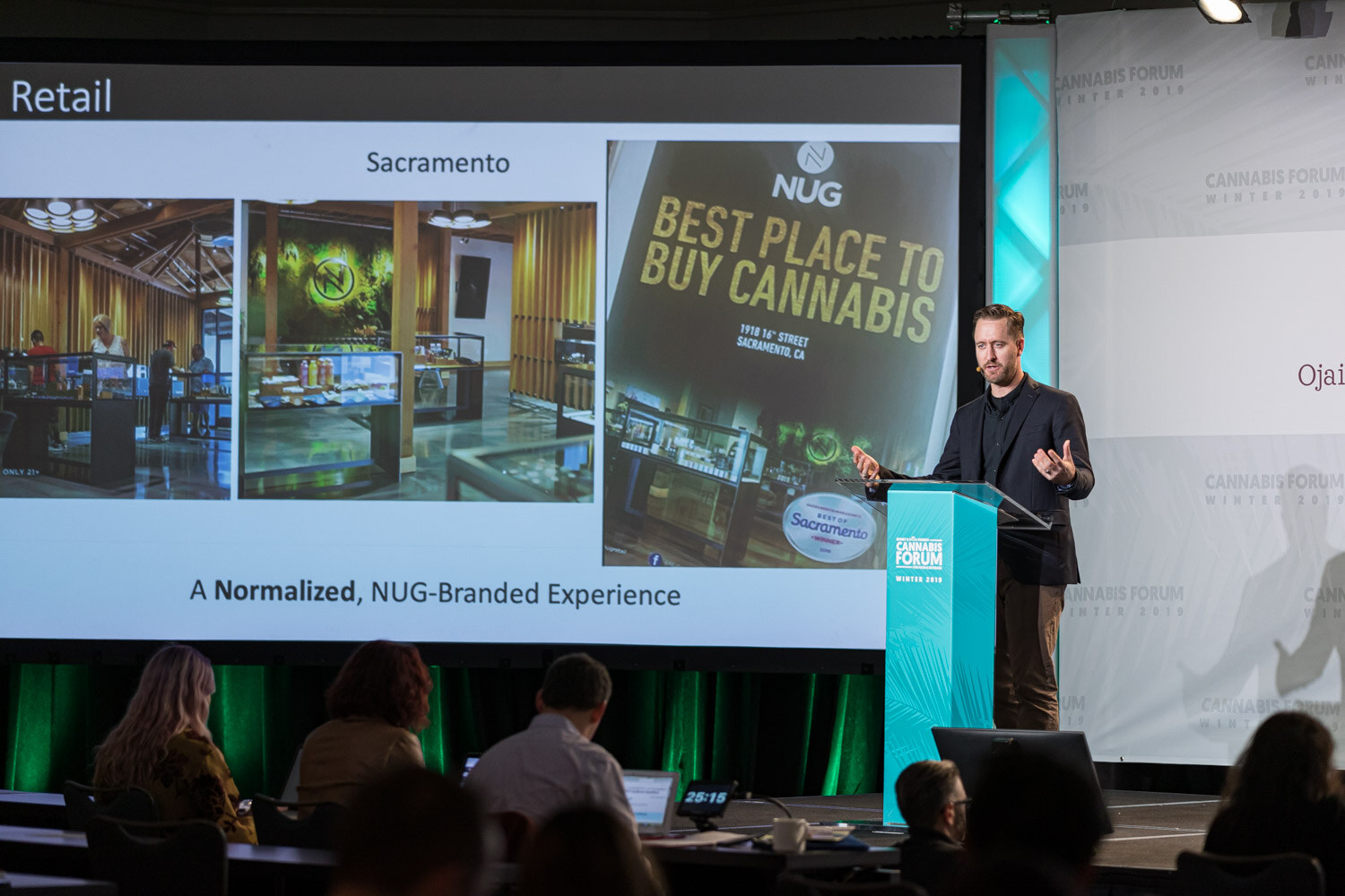 A man in a dark blazer stands at the podium next to a projected retail slide with the NUG logo, store interiors and the caption A Normalized, NUG-Branded Experience.