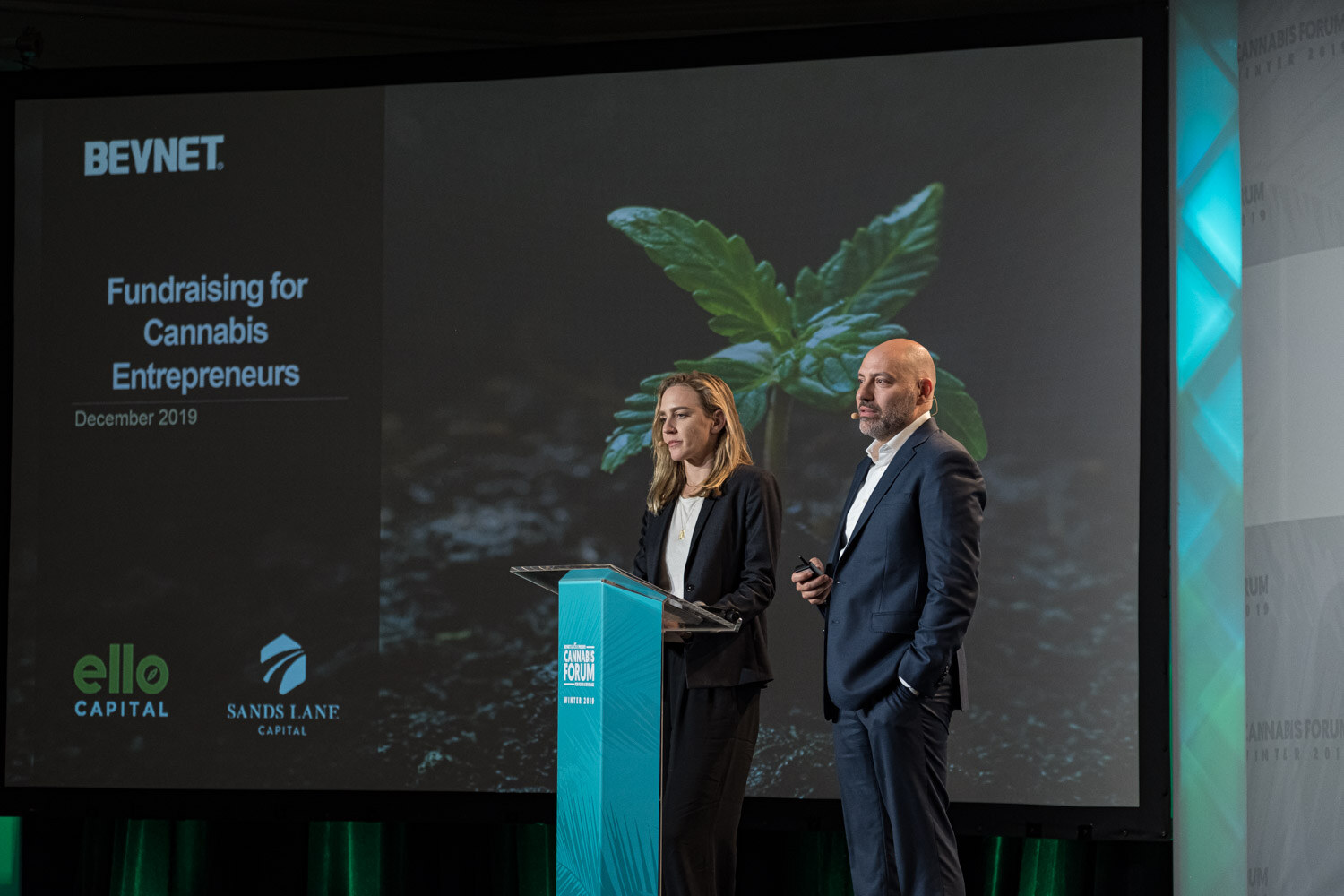 A woman and a man in dark suits stand at a podium beside a slide reading BevNET, Fundraising for Cannabis Entrepreneurs, December 2019 with Ello Capital and Sands Lane Capital logos.