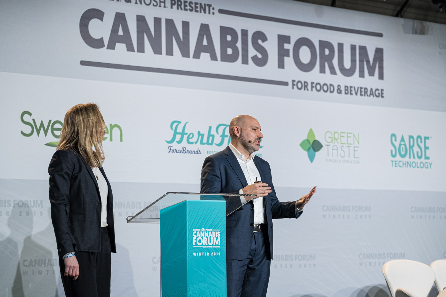 A bald man in a navy suit gestures at the podium while a woman in a dark blazer stands beside him, with Sweegen, HerbForce, Green Taste and Sorse logos behind them.