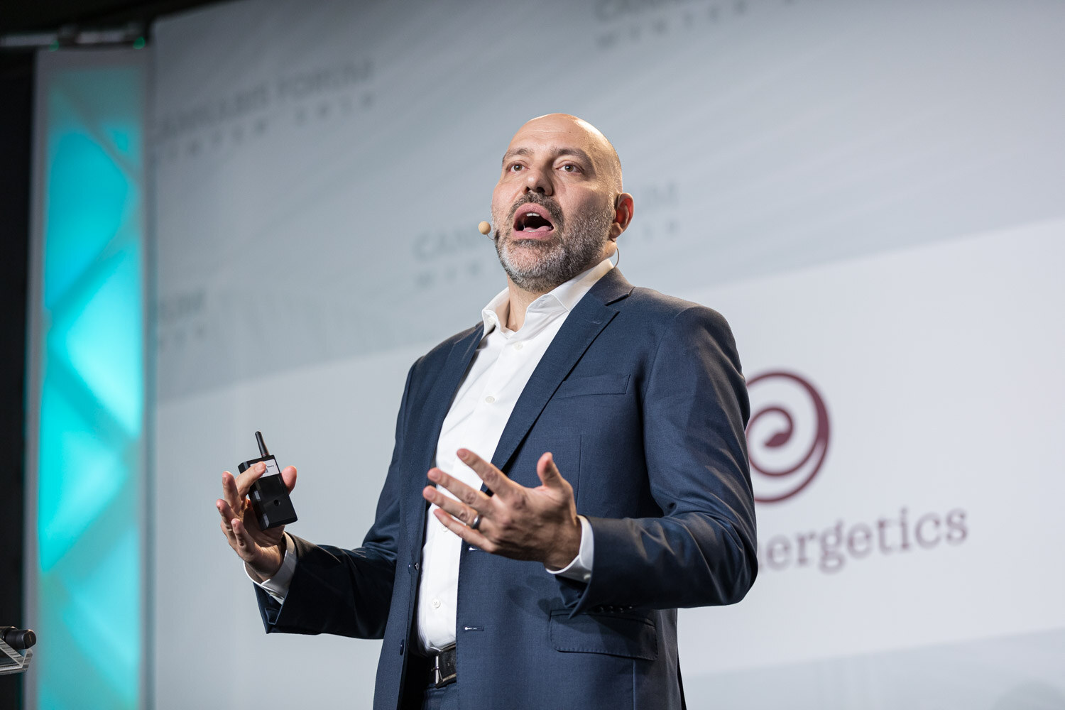 A bald, bearded man in a navy suit speaks with open hands and holds a presentation clicker, in front of a partial Ojai Energetics logo.