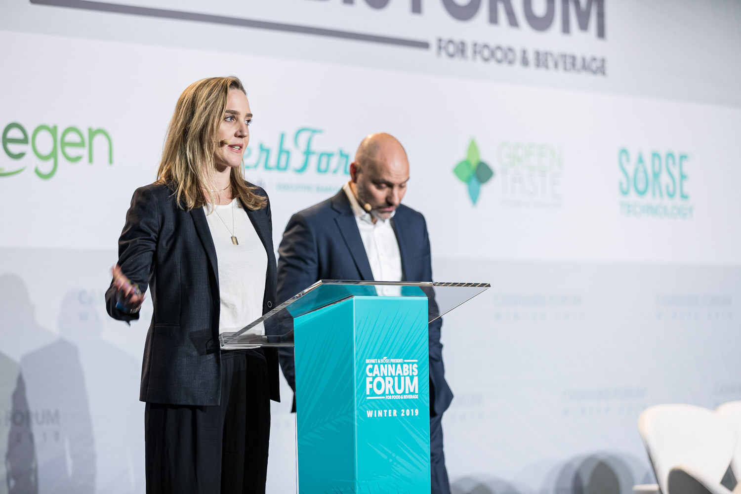 A woman in a dark blazer gestures with an open hand while a bald man in a navy suit stands behind her at a teal Cannabis Forum podium, with HerbForce, Green Taste and Sorse logos behind them.