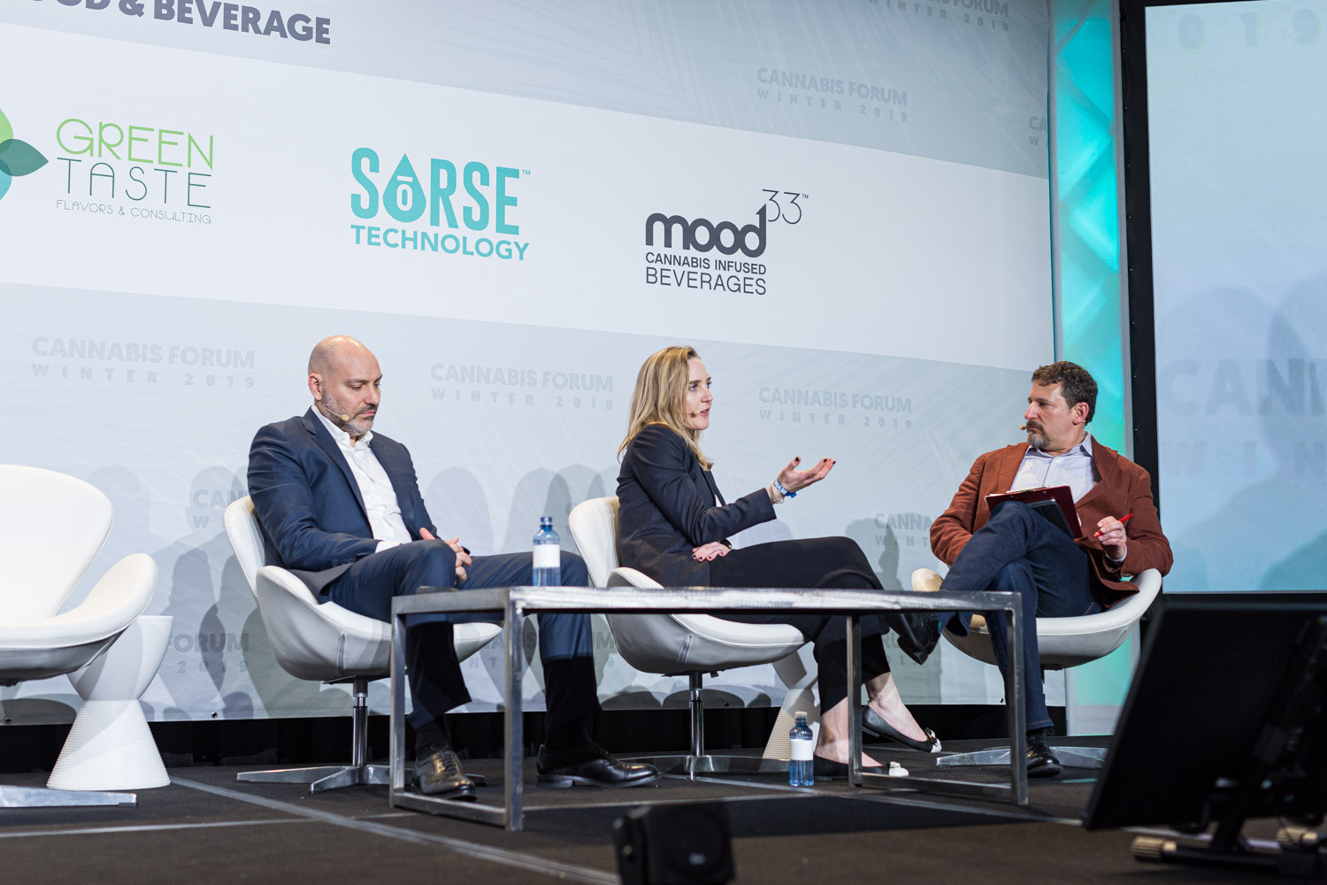 Three people sit in white lounge chairs on stage, a man in a navy suit, a woman in a dark blazer and a man in a rust jacket holding notes, in front of Green Taste, Sorse and Mood33 logos.