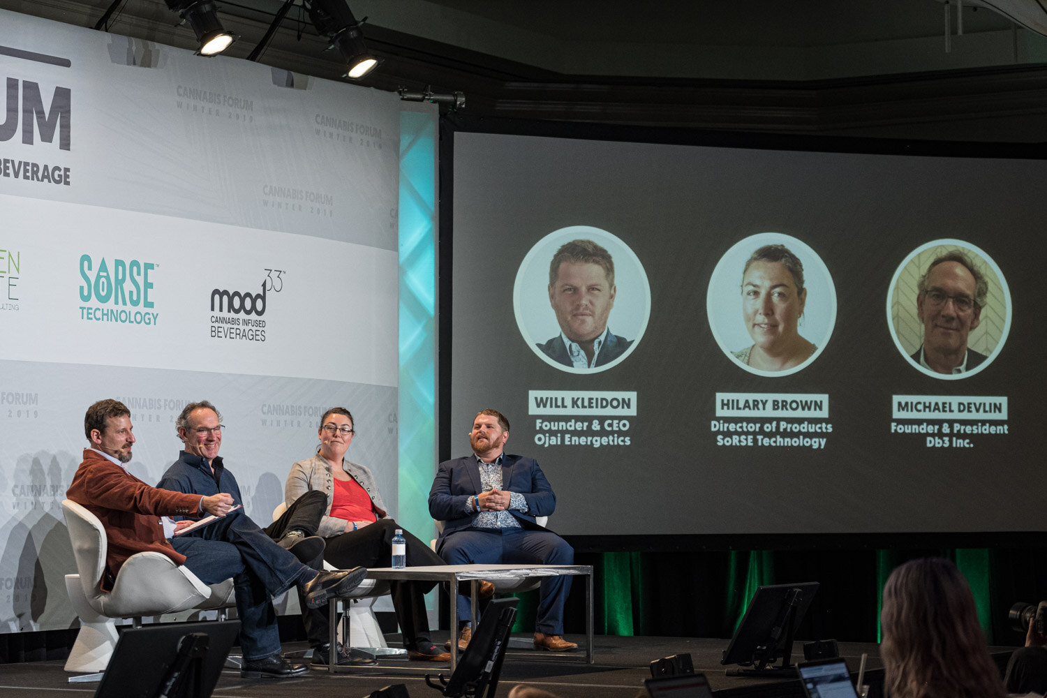 Four people sit on a stage in white lounge chairs under a screen showing three circular portraits with their names and company titles for Ojai Energetics, Sorse Technology and Db3 Inc.