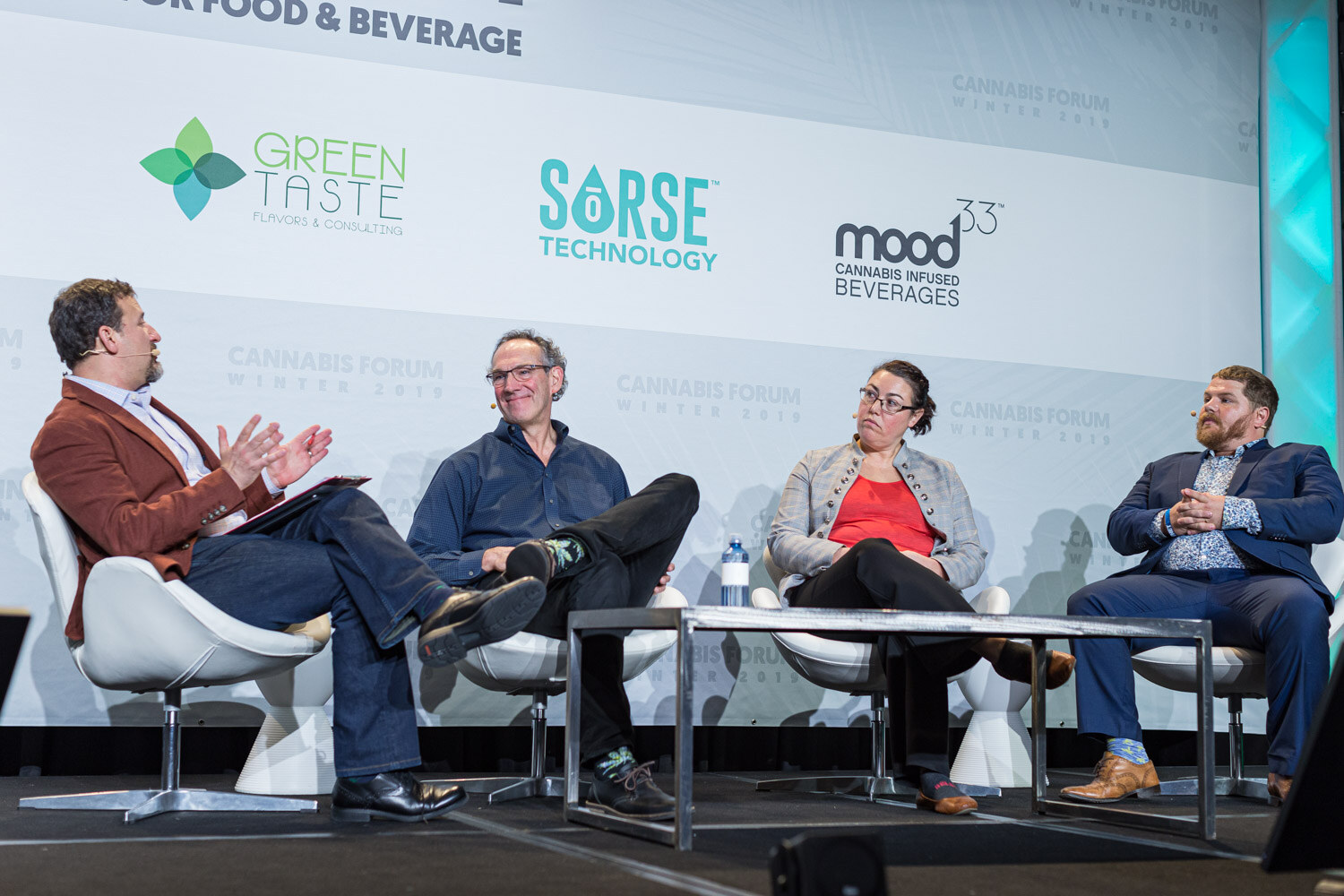 Four people sit on a stage in white chairs, a man in a rust jacket with a clipboard talking with a man in a blue shirt, a woman in a gray jacket and a man in a navy jacket, in front of Green Taste, Sorse and Mood33 logos.