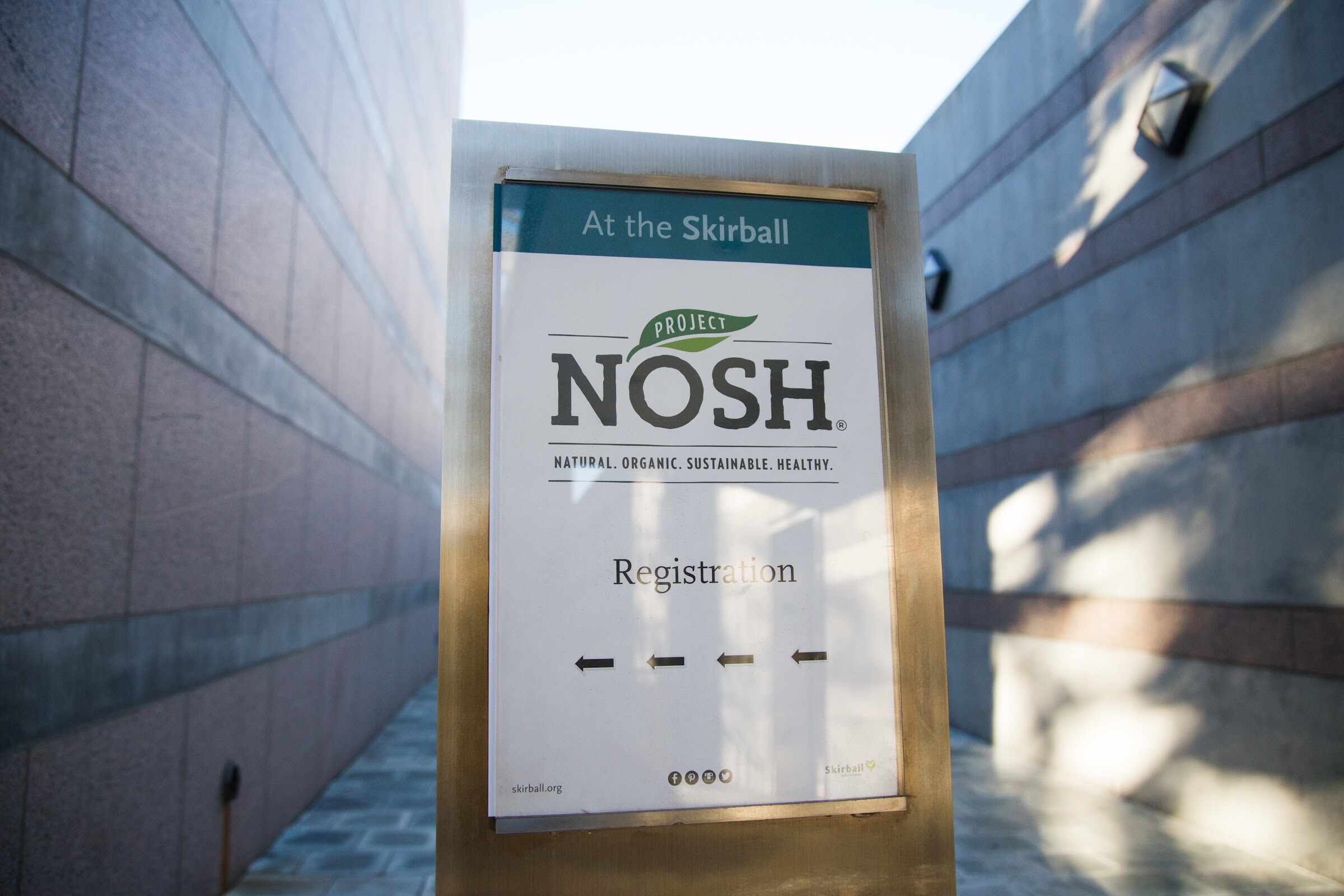 A framed sign between two stone walls reads At the Skirball, Project NOSH, Registration, with four arrows pointing left.