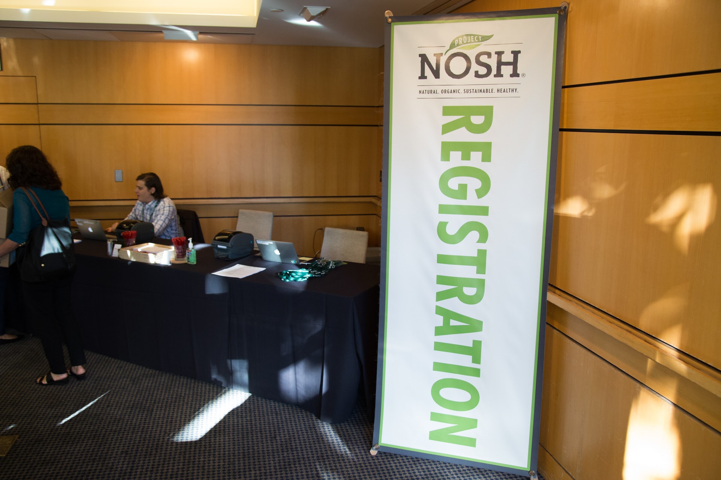 A man sits at a black-draped table with laptops and a label printer while an attendee stands at left; a tall Project NOSH Registration banner stands to the right.