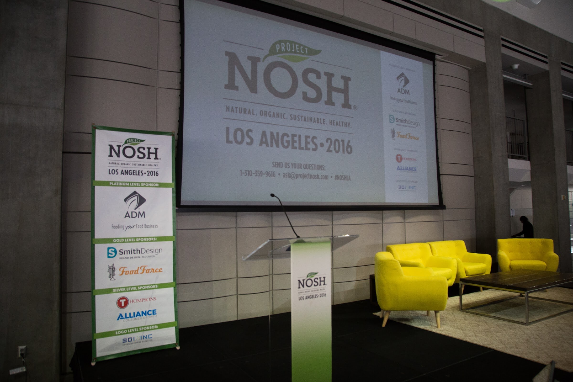 An empty stage with a clear lectern, three yellow armchairs and a coffee table beside a projection screen reading Project NOSH Los Angeles 2016 and a tall sponsor banner.
