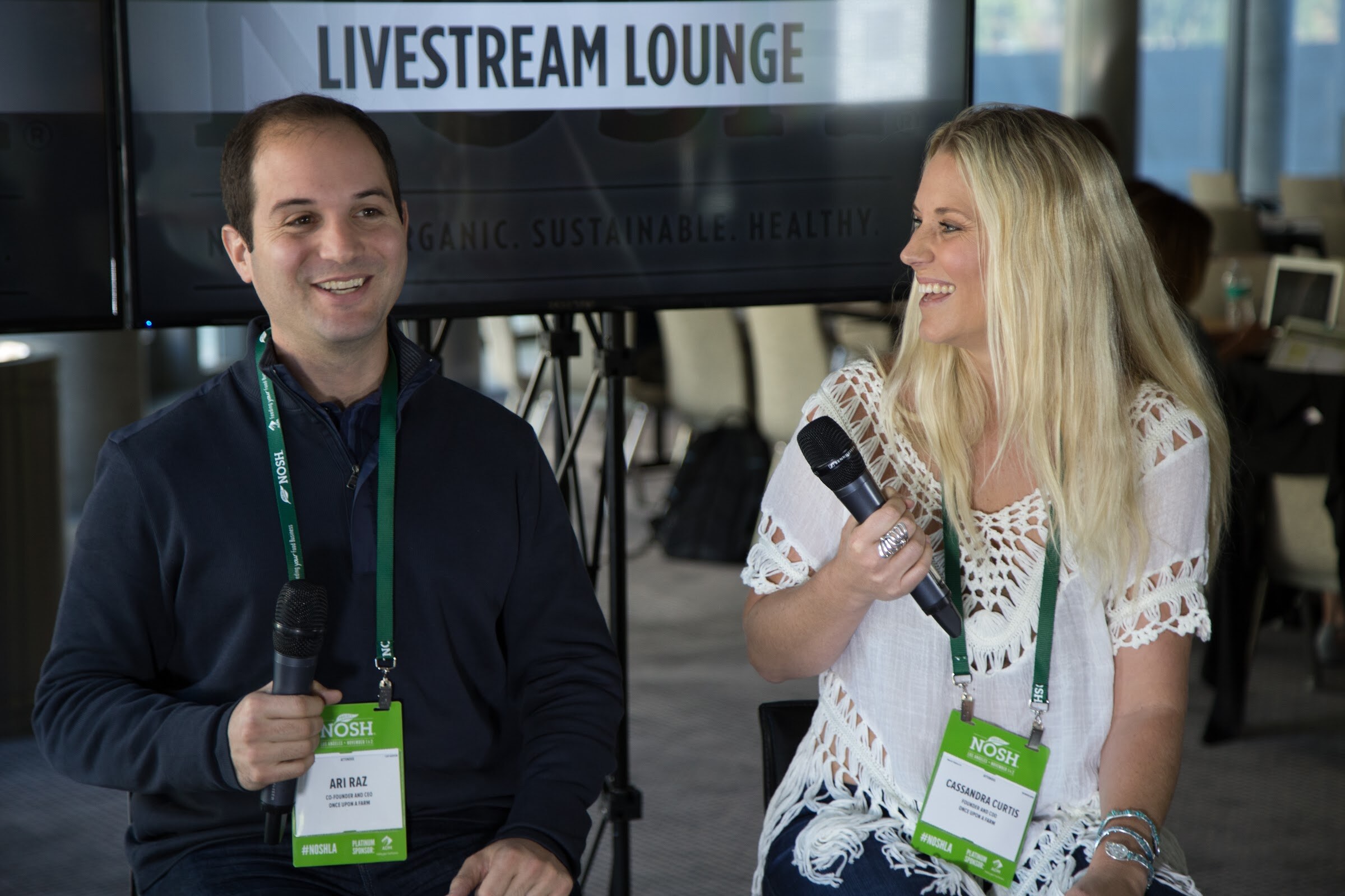 A man in a navy zip pullover and a woman with long blonde hair in a white crochet top hold microphones and smile in front of a monitor reading Livestream Lounge.