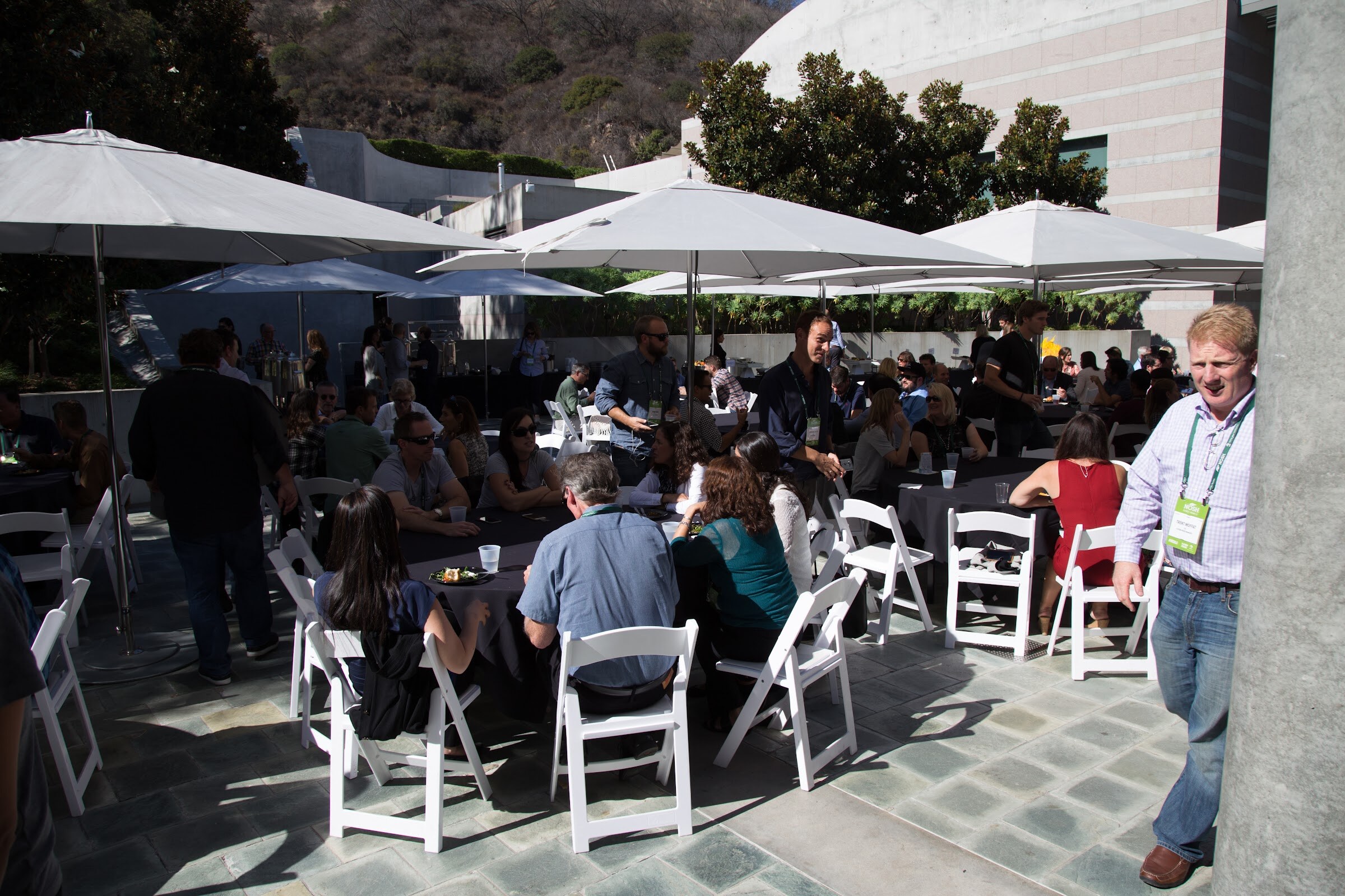Groups of people sit and stand at tables with white folding chairs under large white umbrellas on a sunlit stone patio.
