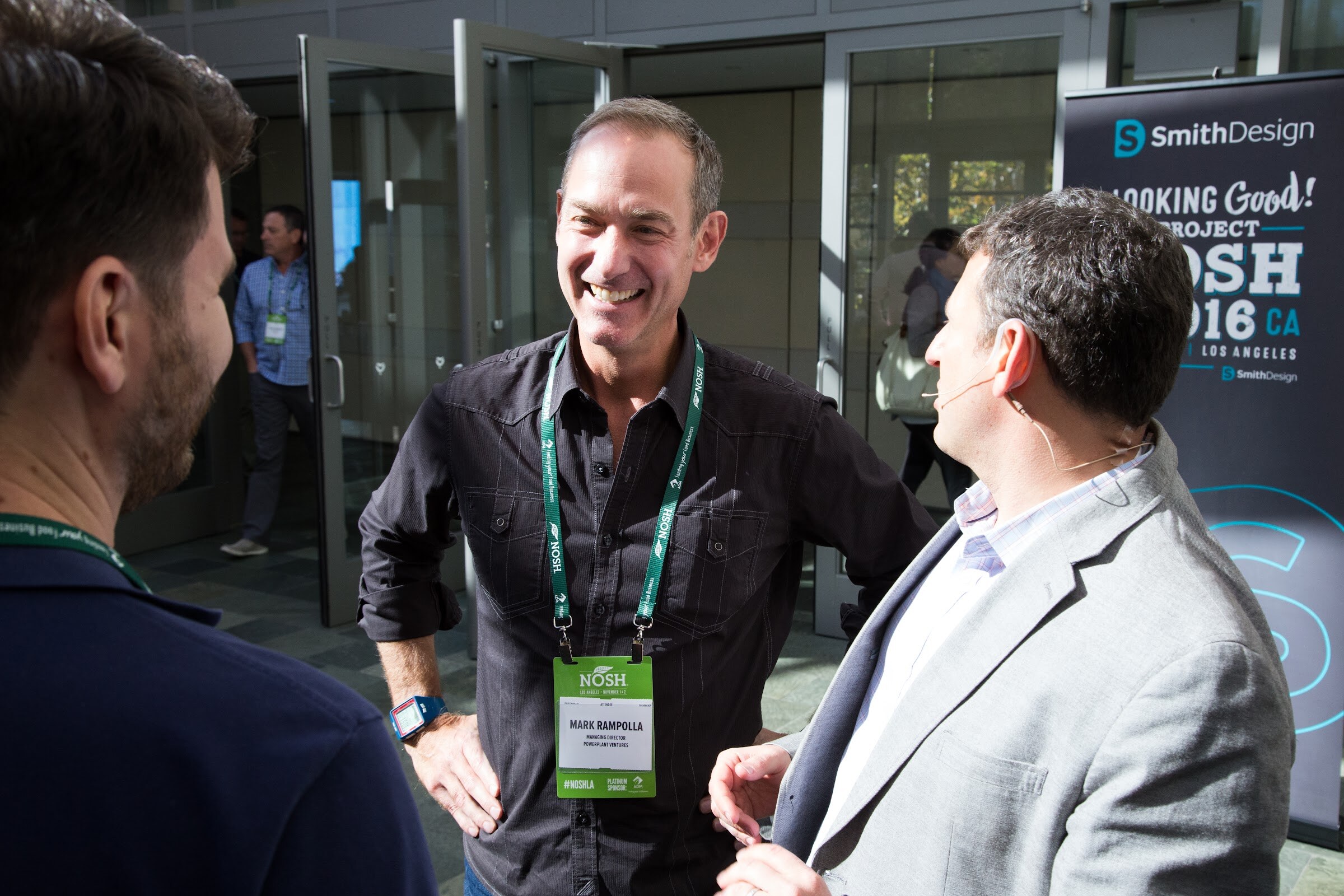 A smiling man in a dark shirt with a NOSH badge talks with two other men beside a black SmithDesign banner reading Looking Good at Project NOSH 2016 CA.