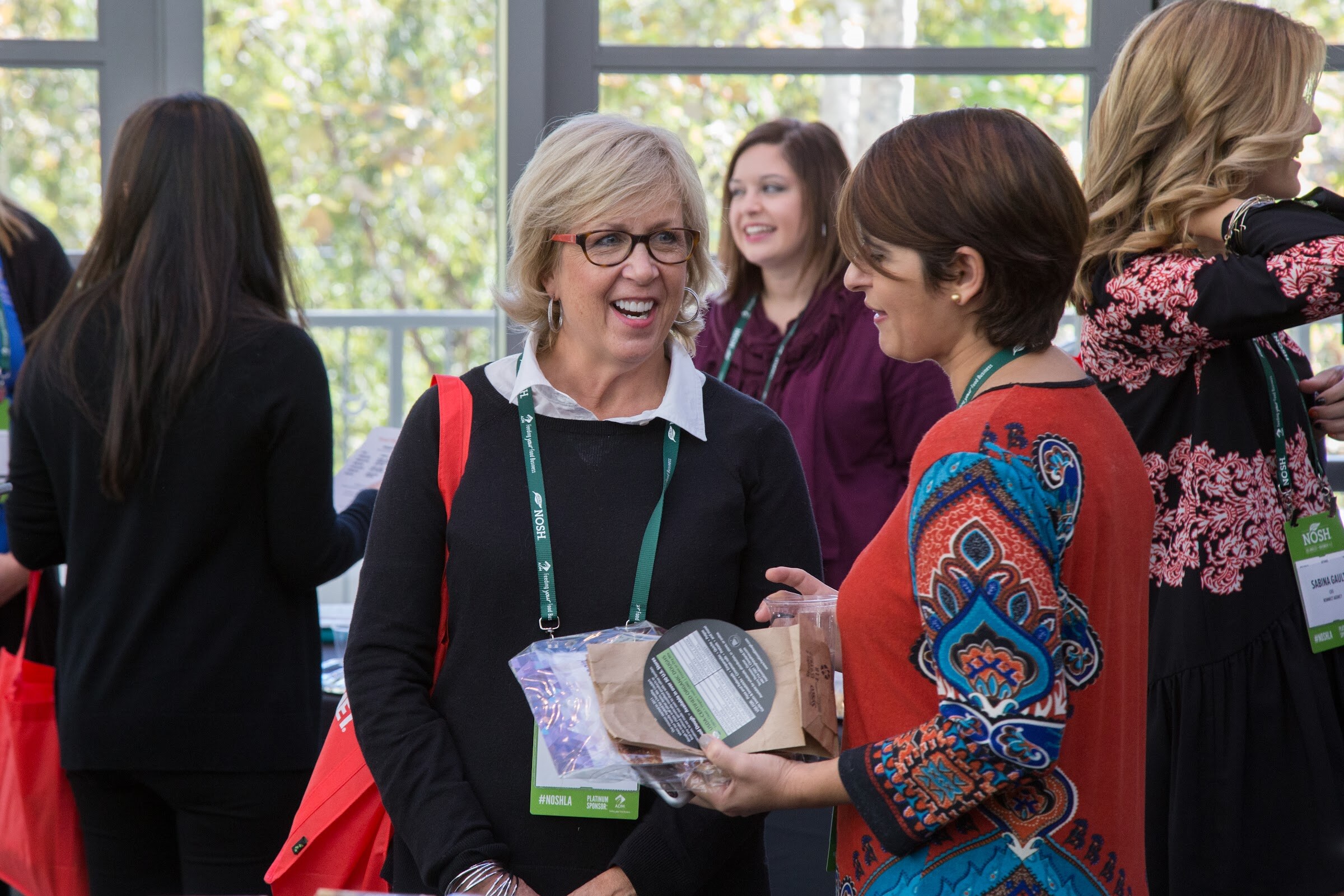 A smiling woman with short blonde hair and glasses holds a paper bag and a plastic bag of samples while talking with a woman in a red patterned top; both wear NOSH lanyards.