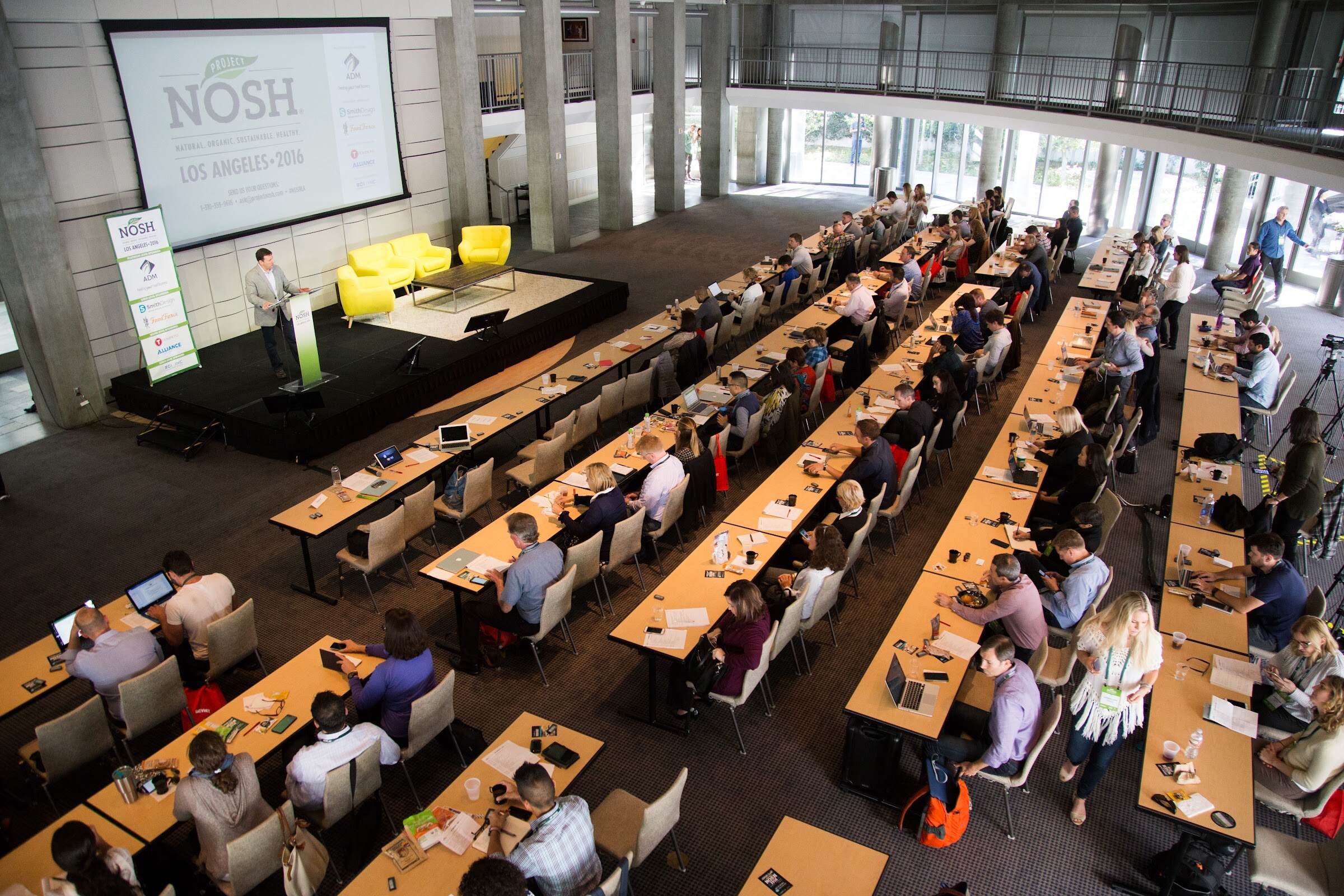 High-angle view of a ballroom with a speaker at a lectern on a stage with yellow armchairs, a NOSH projection screen, two green NOSH logos projected on the wall, and rows of attendees at long tables.