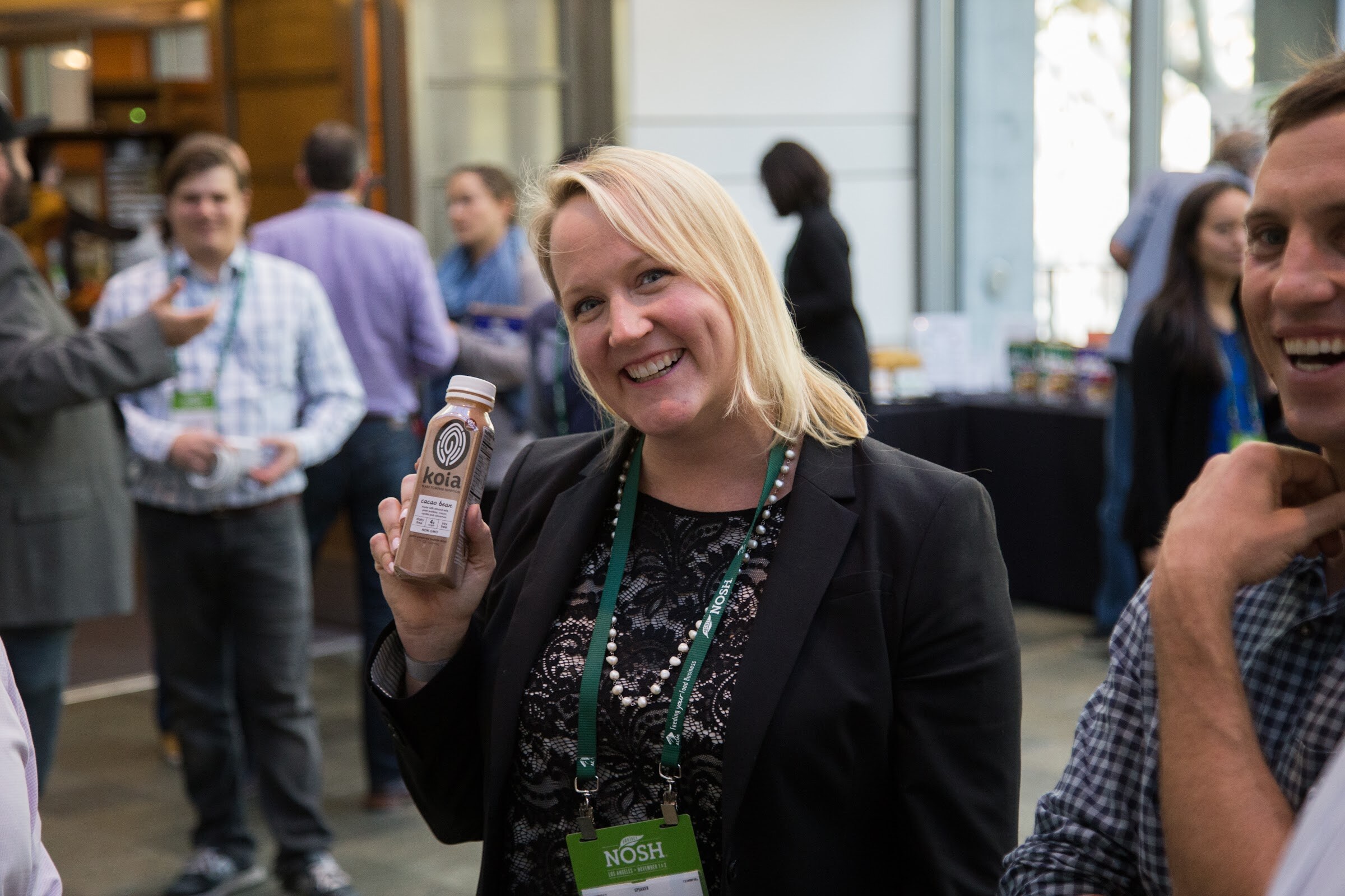 A smiling woman with blonde hair and a NOSH lanyard holds up a bottle of Koia drink with the room full of attendees blurred behind her.