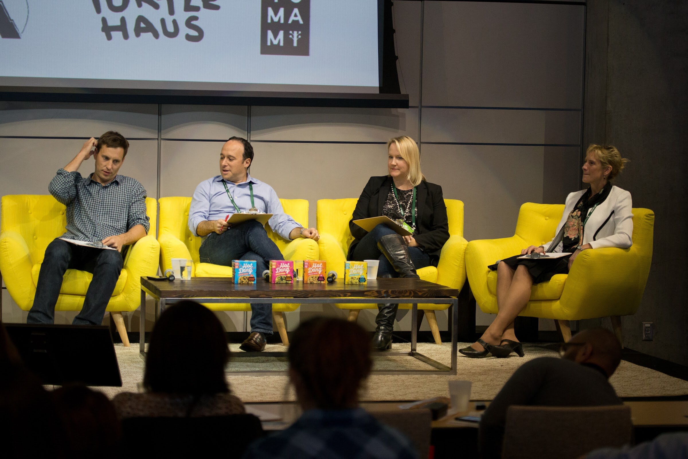 Two men and two women on yellow armchairs on a stage, each holding a clipboard, with several Hot Dang breakfast toaster boxes lined up on the coffee table and a slide with Turtle Haus and Yumami logos above.