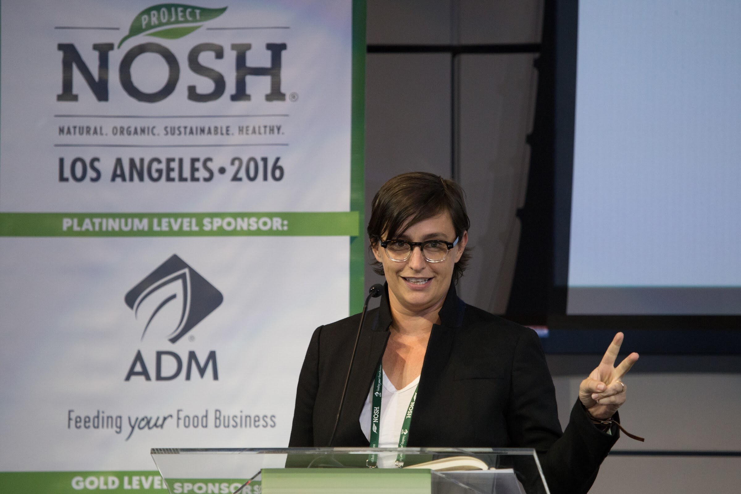 A woman with short dark hair and black-framed glasses stands at a lectern with a microphone and raises two fingers; the Project NOSH Los Angeles 2016 banner with ADM behind her.