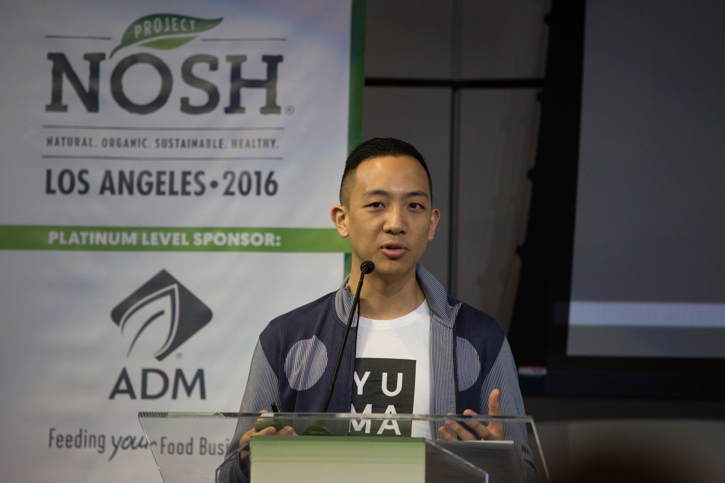 A man in a navy zip jacket over a white Yumami t-shirt speaks at a clear lectern with a microphone, with the Project NOSH Los Angeles 2016 and ADM banner behind him.