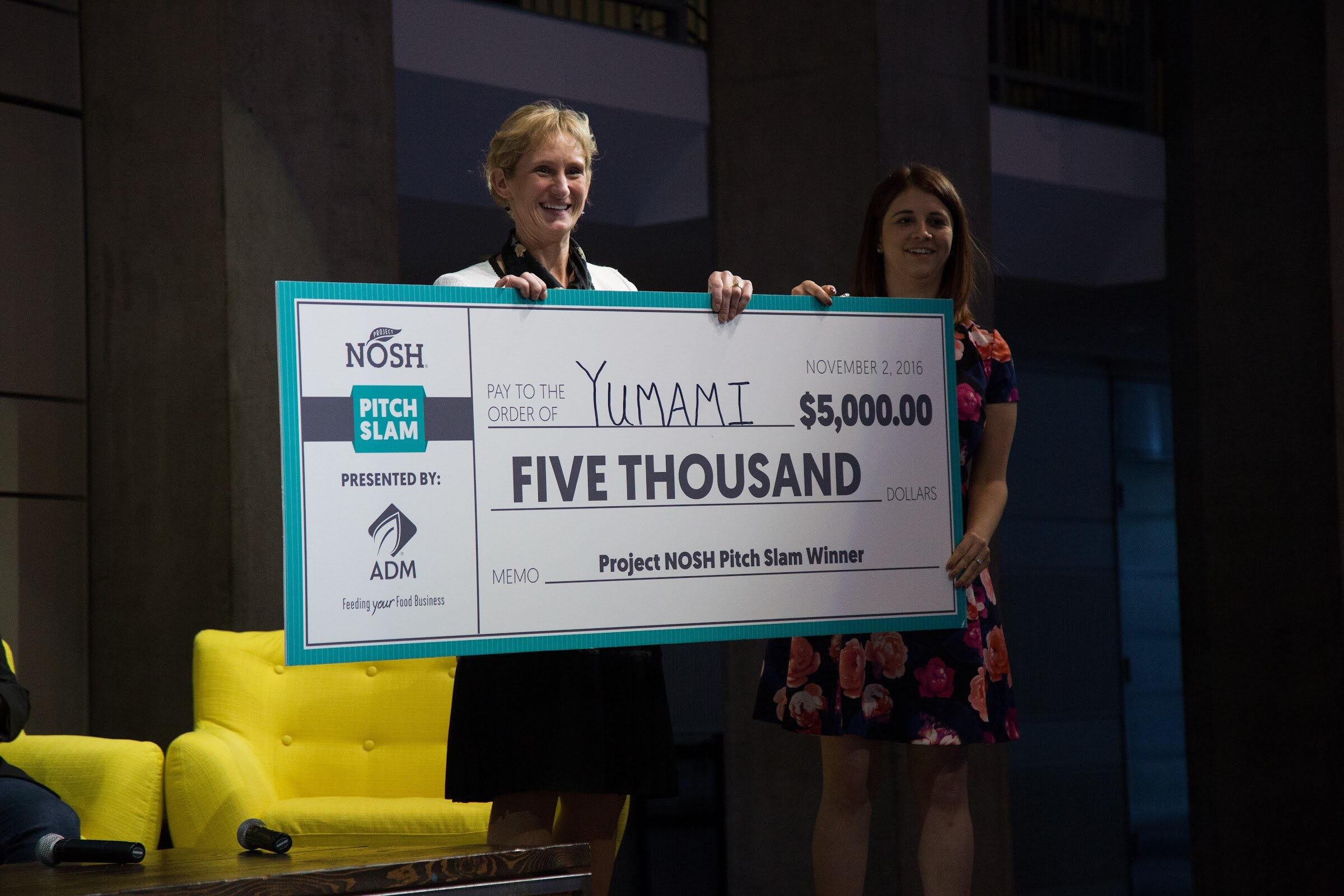 Two women on stage hold a large teal and white check reading Pay to the order of Yumami, five thousand dollars, Project NOSH Pitch Slam Winner, presented by ADM, dated November 2, 2016.
