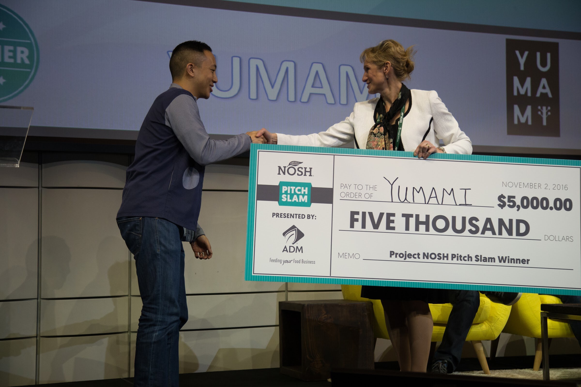 On stage, a man in a navy vest and jeans shakes hands with a woman in a white blazer who holds a large check for $5,000 made out to Yumami; the slide behind reads Yumami.