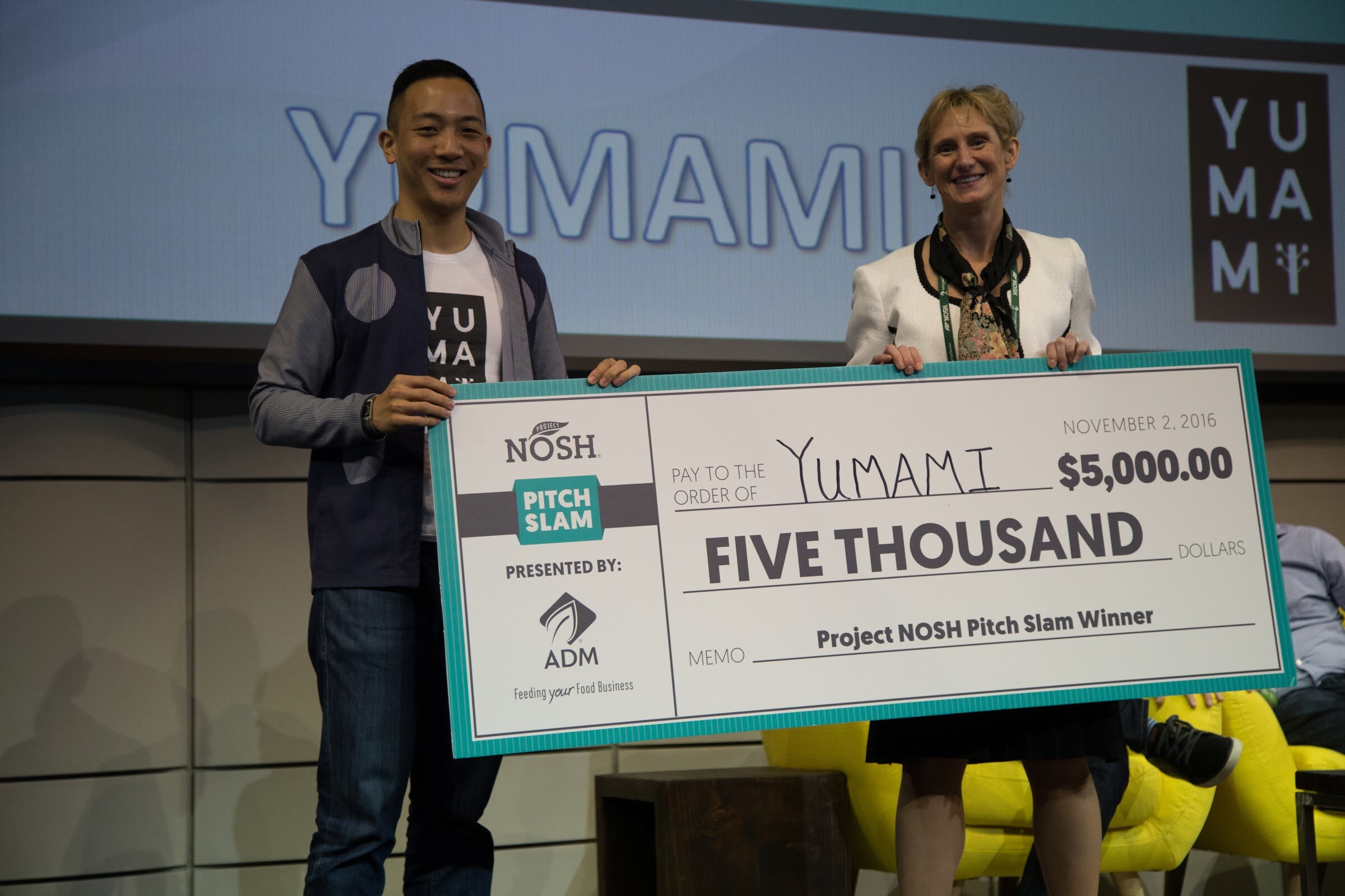 A smiling man in a navy vest and white Yumami t-shirt and a smiling woman in a white jacket hold a large teal check for $5,000 payable to Yumami on a stage with a Yumami slide behind them.