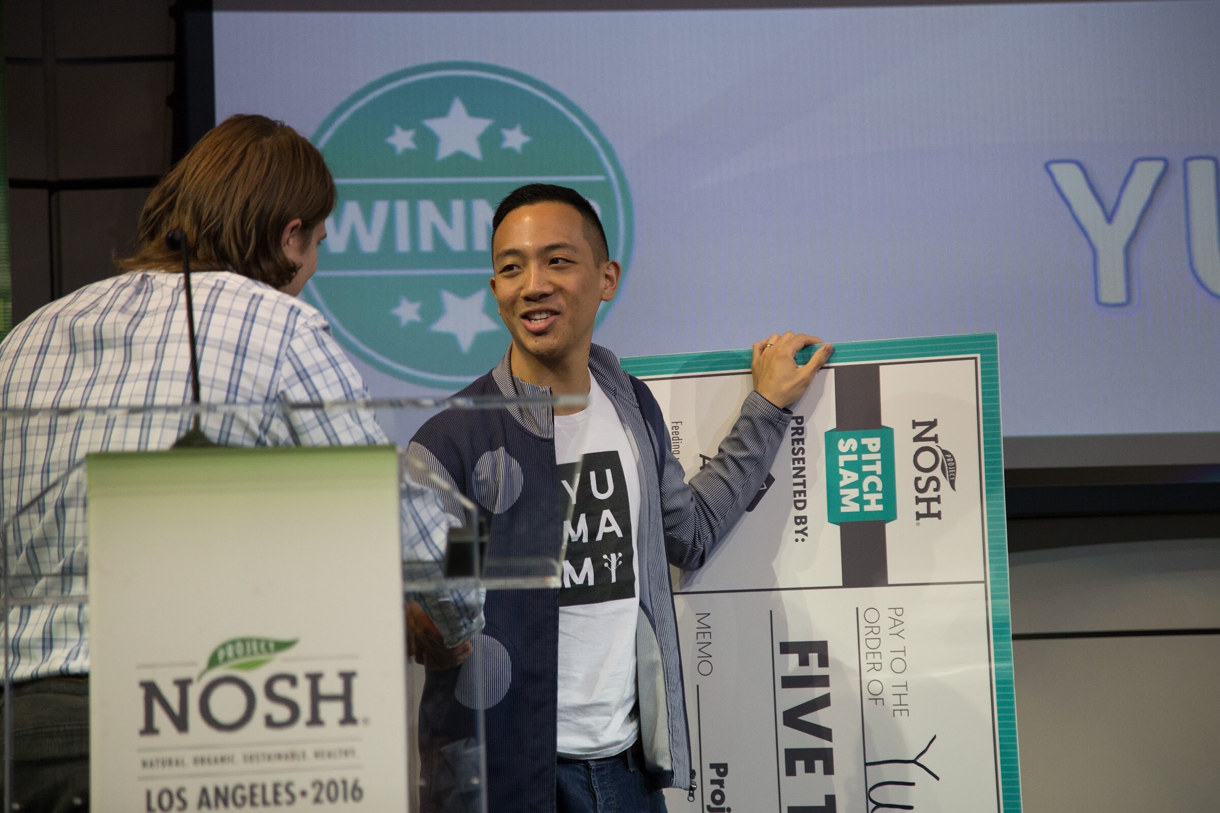 A smiling man in a navy jacket and white Yumami t-shirt stands beside a giant Pitch Slam check, with a Winner badge on the projected slide and the NOSH lectern in the foreground.