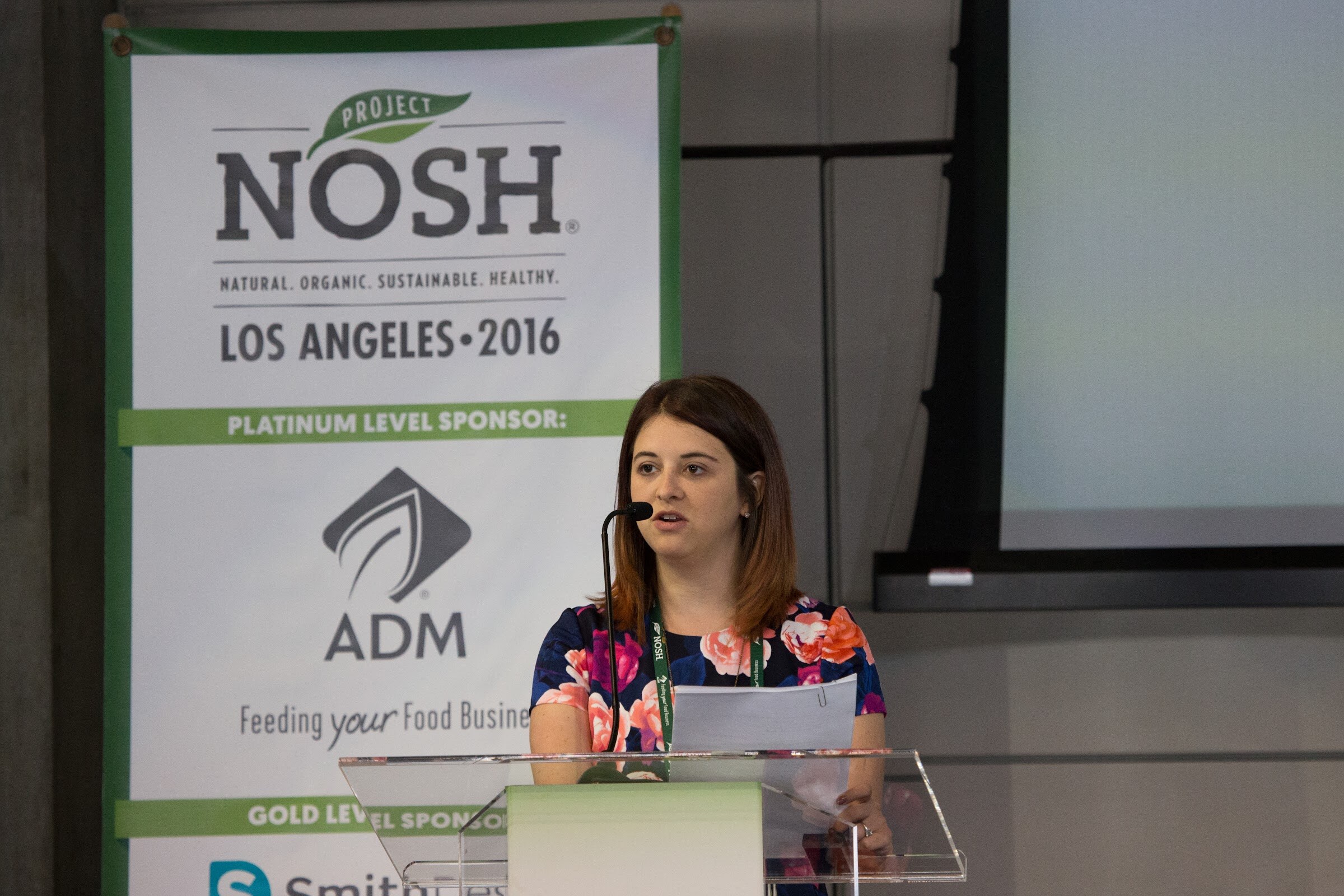A woman in a bright floral dress speaks into a microphone at a clear lectern, with a Project NOSH Los Angeles 2016 banner listing ADM as platinum level sponsor.