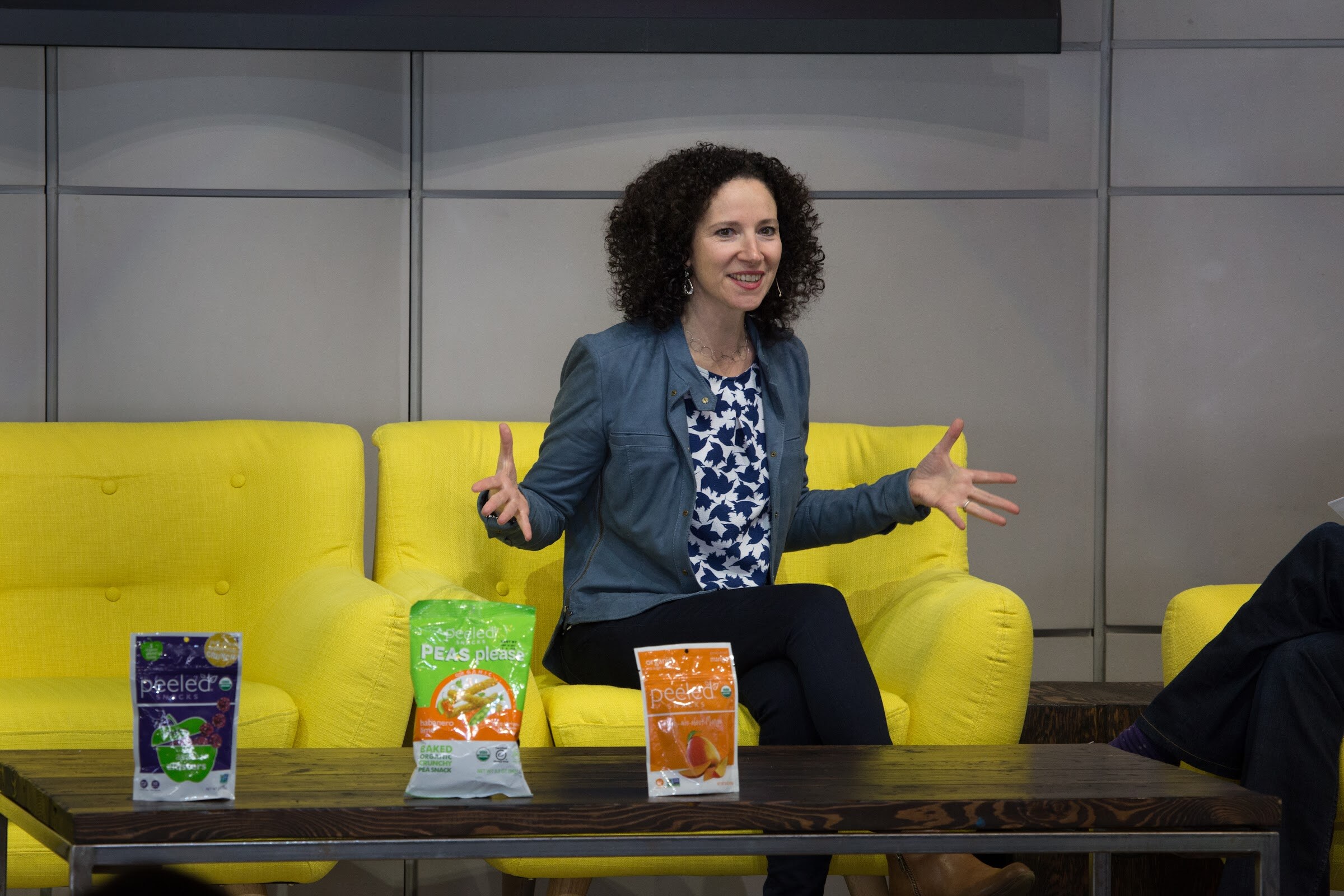 A smiling woman in a blue jacket with her hands held out sits on a yellow sofa; three Peeled Snacks bags stand on the low wooden table in front of her.