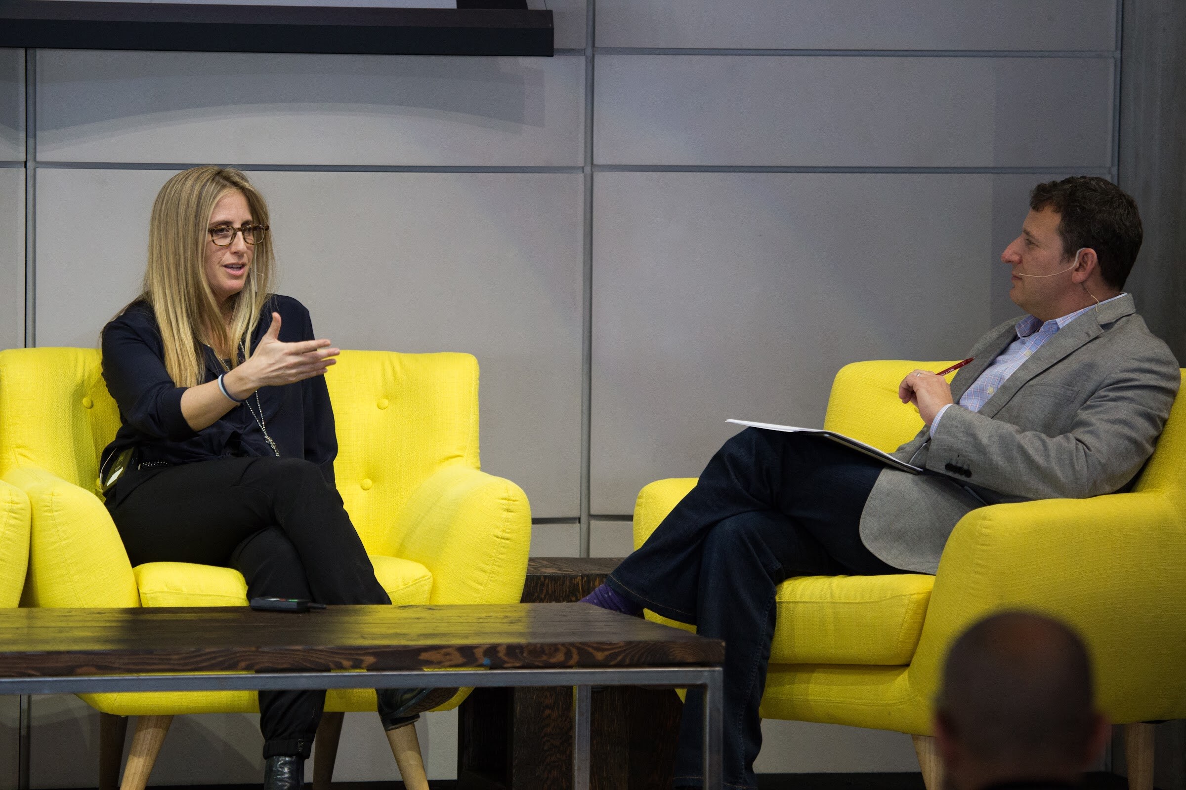 A woman with glasses and long blonde hair gestures as she speaks to a man in a gray blazer who sits across from her with papers and a red pen.