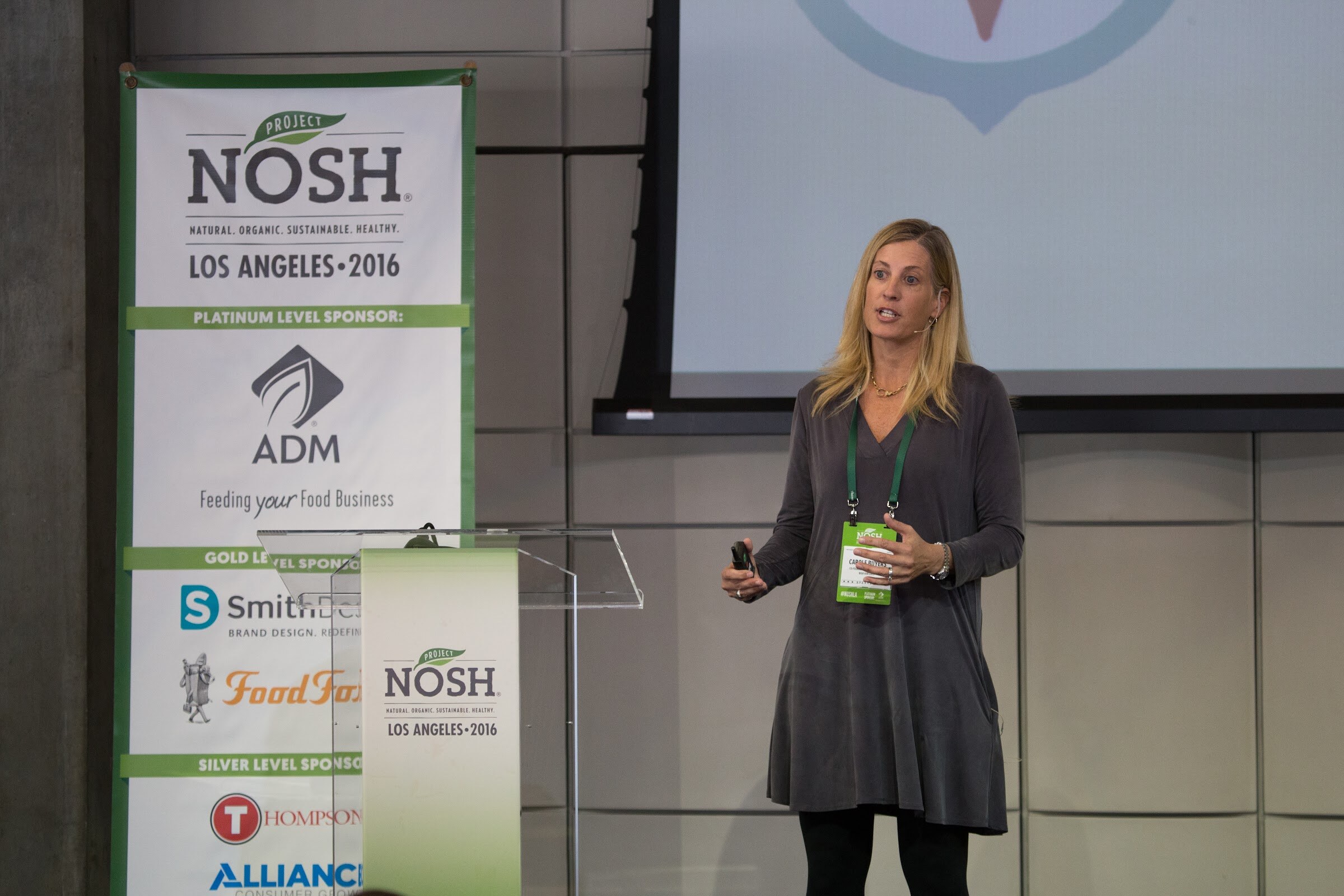 A woman in a gray dress and lanyard speaks into a headset microphone next to a clear lectern; the sponsor banner behind her lists ADM, SmithDesign, Food Force, Thompson and Alliance.