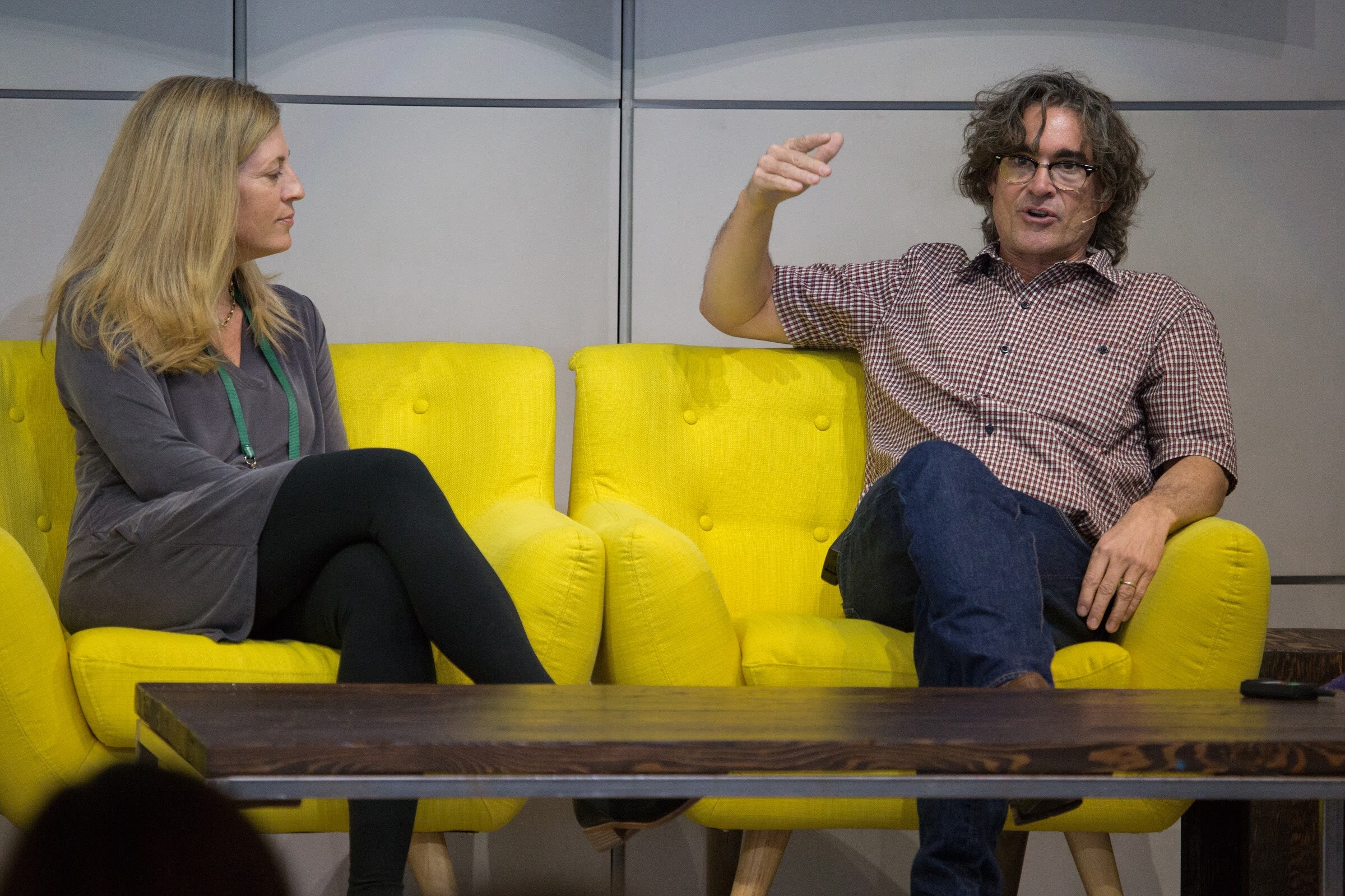 A woman in a gray top listens on a yellow sofa while a man in a red checked shirt and jeans gestures with one arm over the sofa back.
