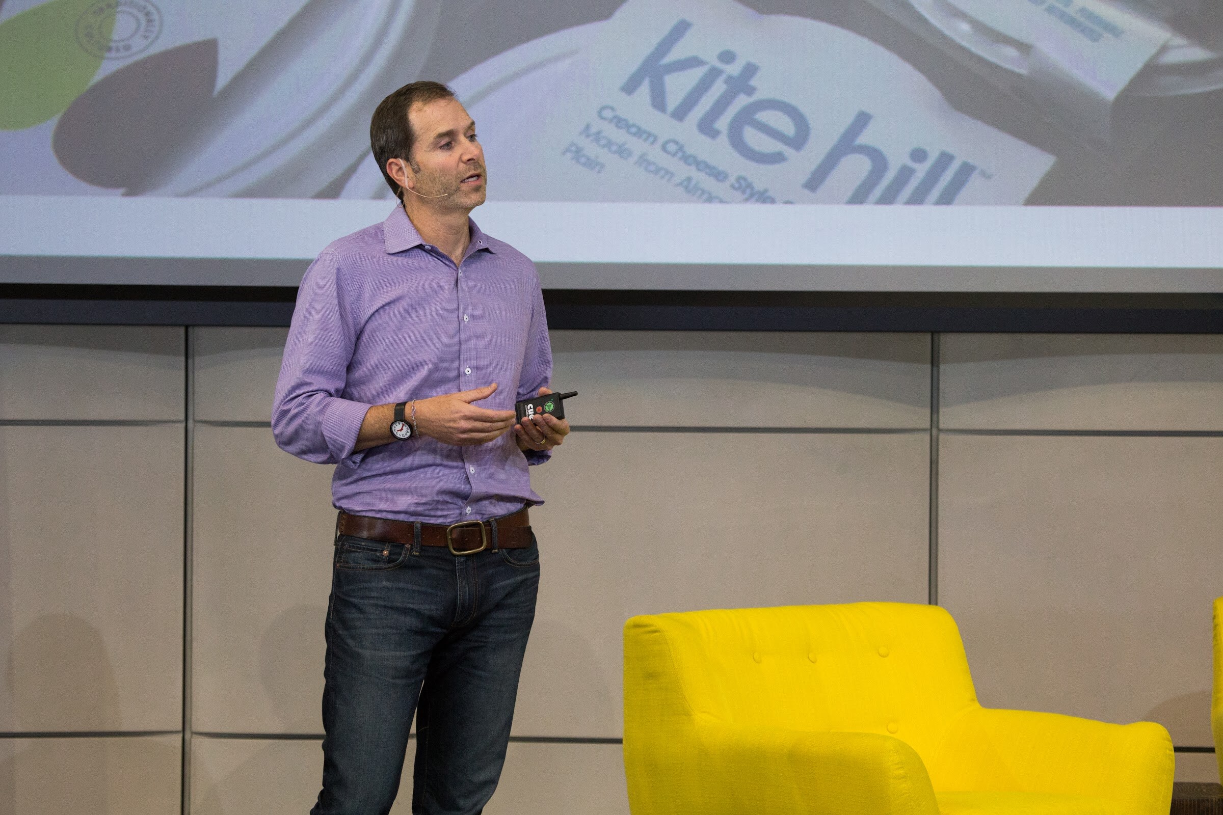 A man in a lavender shirt and jeans holds a clicker and speaks while standing on stage; the slide above him shows Kite Hill cream cheese style spread.