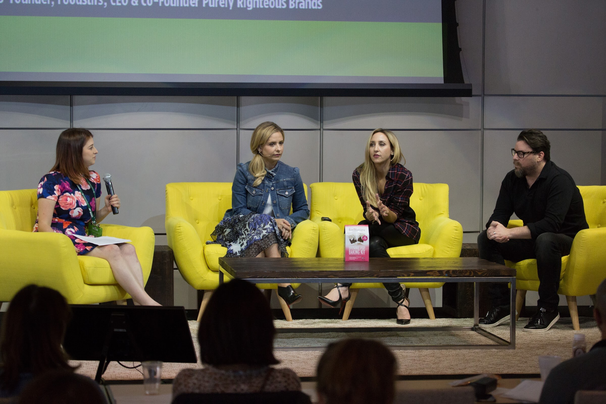 Four people on yellow chairs on a stage; a woman in a floral dress holds a microphone, and a pink Foodstirs baking kit box stands on the coffee table between them.